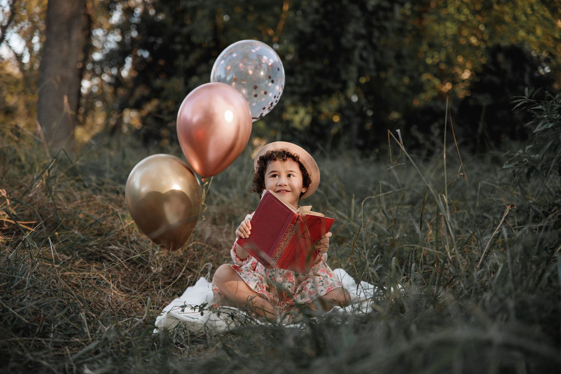 Una niña está sentada en el césped sosteniendo globos y leyendo un libro.