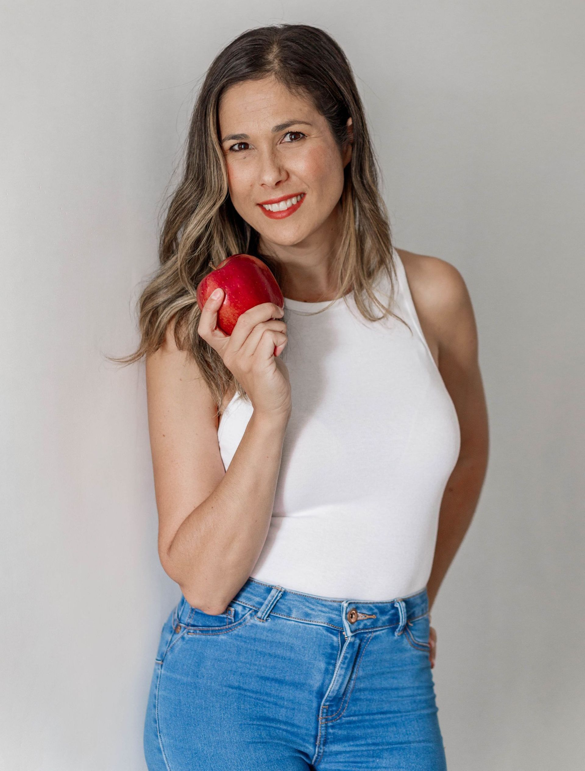 Una mujer con una camiseta blanca y vaqueros azules sostiene una manzana roja.