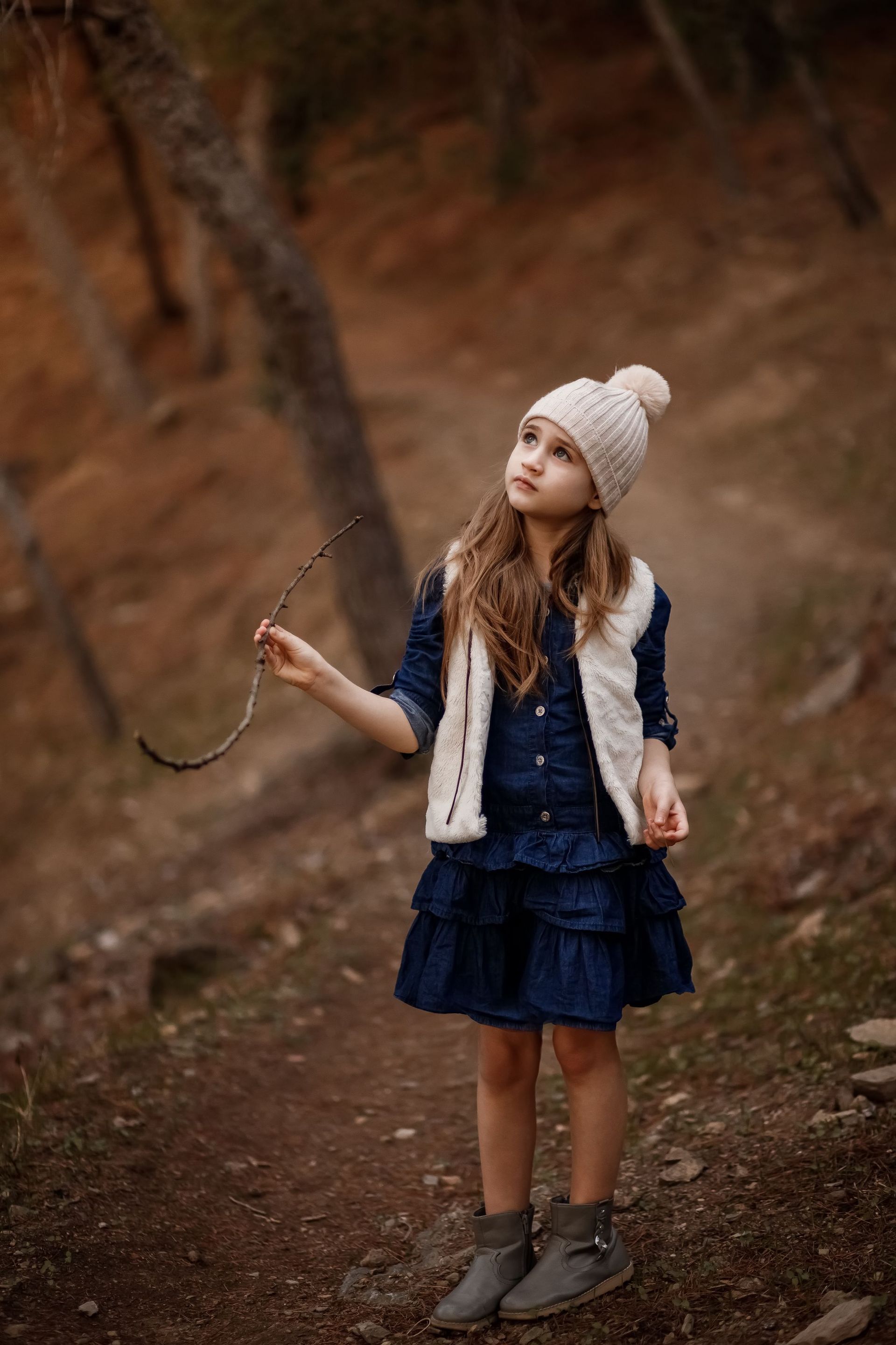 Una chica con sombrero y chaleco blancos sostiene una ramita y mira hacia arriba. Lleva un vestido azul y botas grises. La escena es al aire libre.