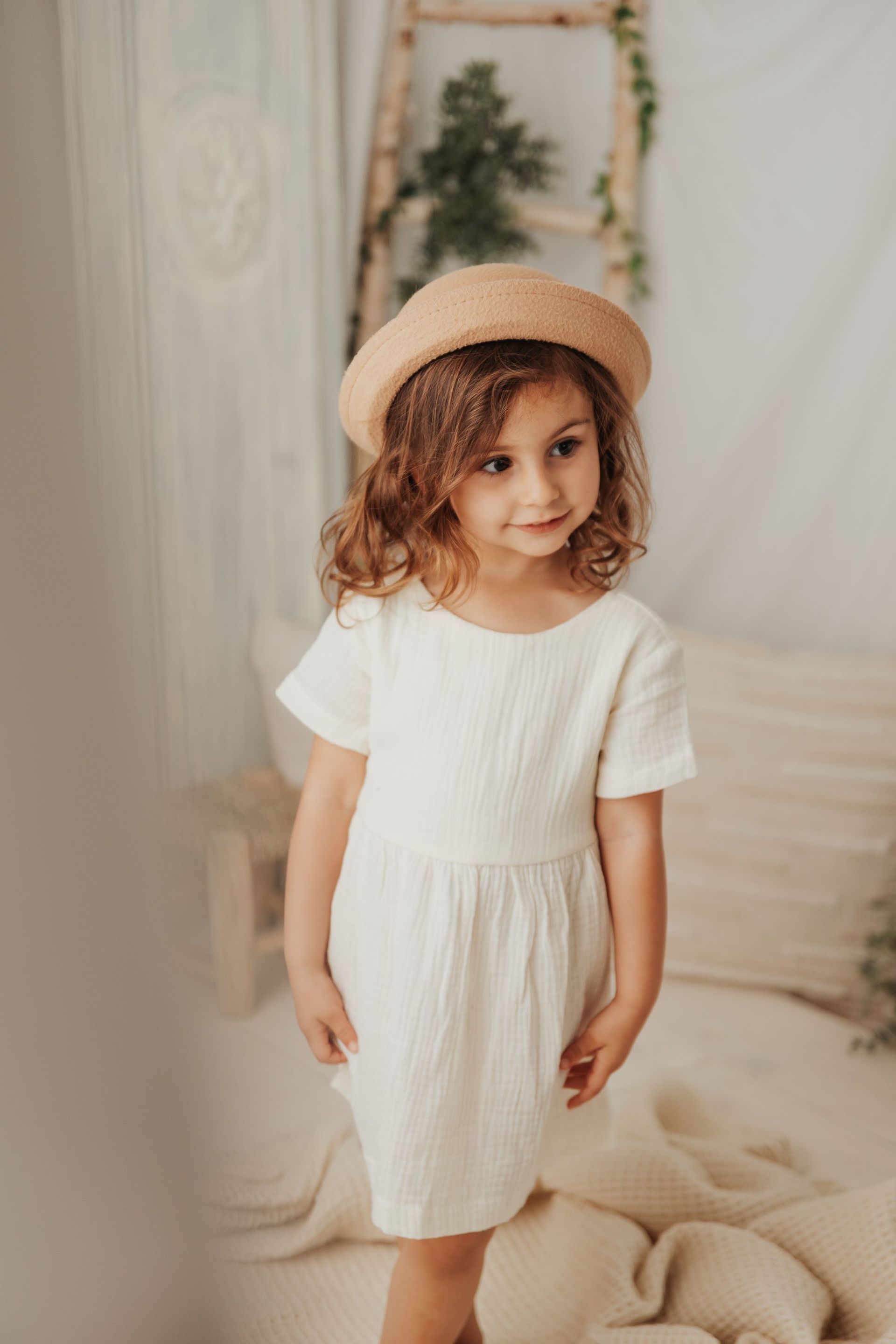 Una niña que lleva un vestido blanco y un sombrero está parada en una cama.