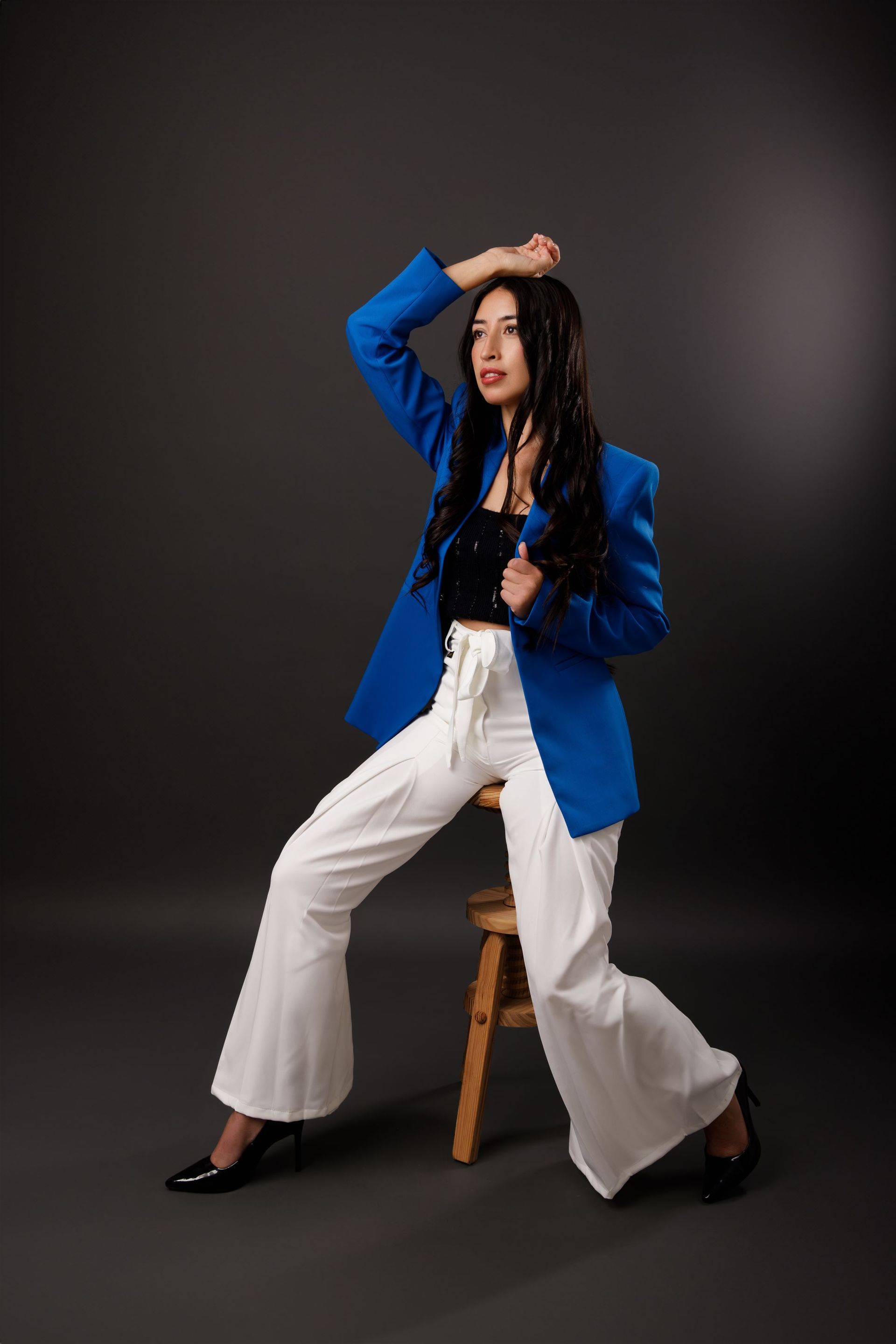 Una mujer con una chaqueta azul y pantalones blancos está sentada en un taburete de madera.