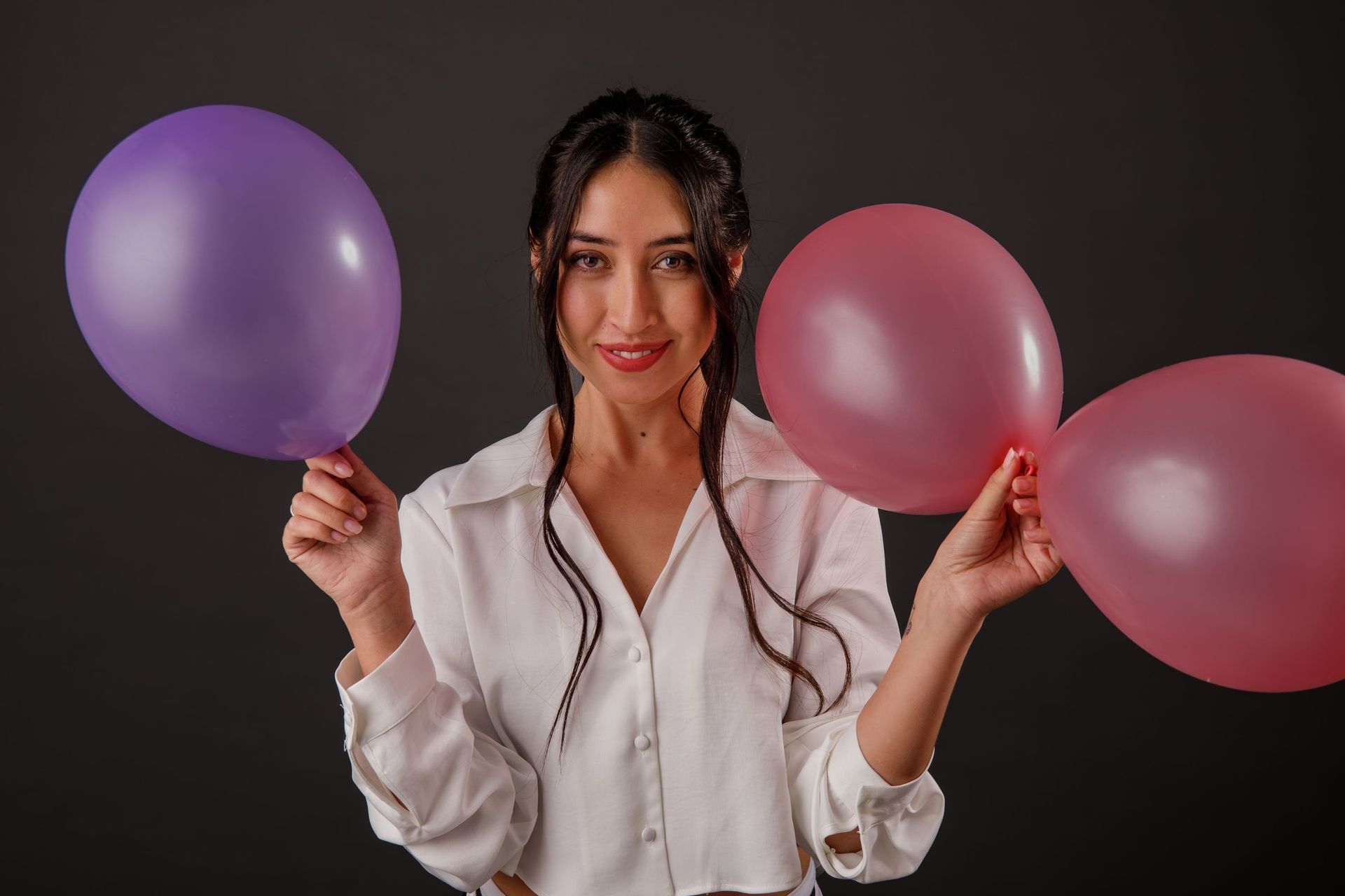 Mujer con camisa blanca sosteniendo globos morados y rosas, sonriendo sobre un fondo oscuro.