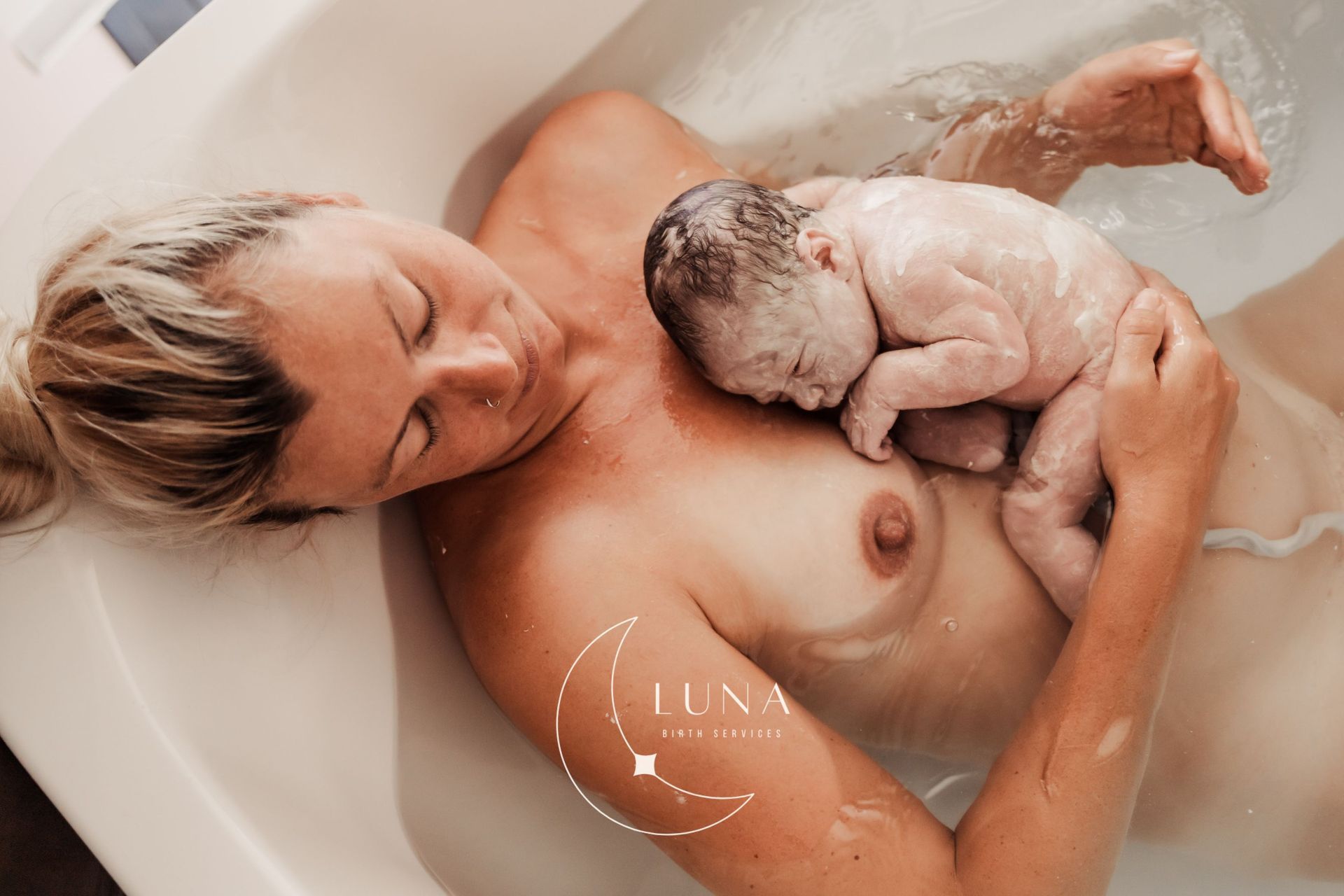Una mujer en la bañera acuna a su bebé recién nacido, ambos sumergidos en el agua. El bebé descansa sobre su pecho.