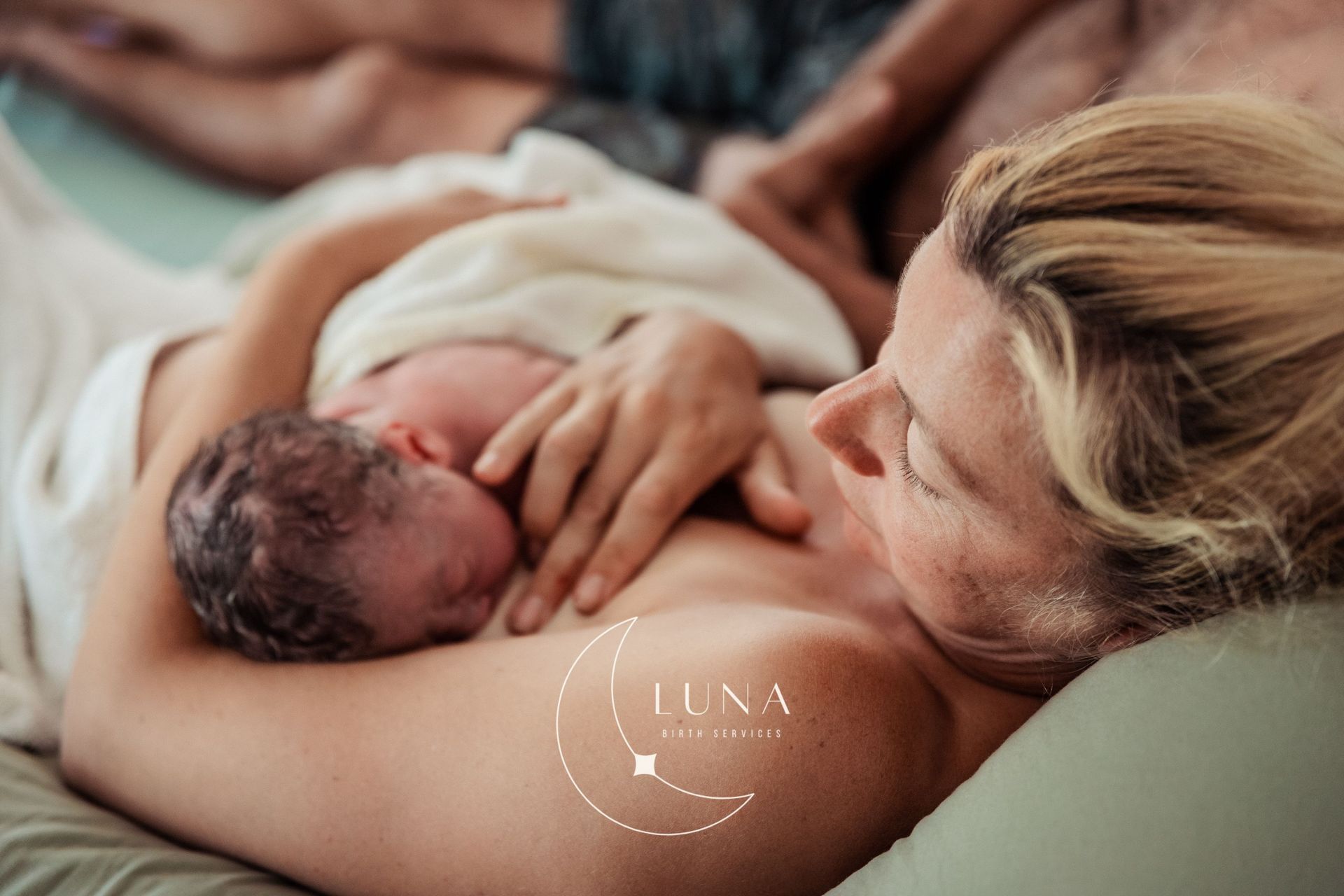 Recién nacido amamantando. La madre sostiene al bebé en contacto piel con piel. Pareja al fondo.