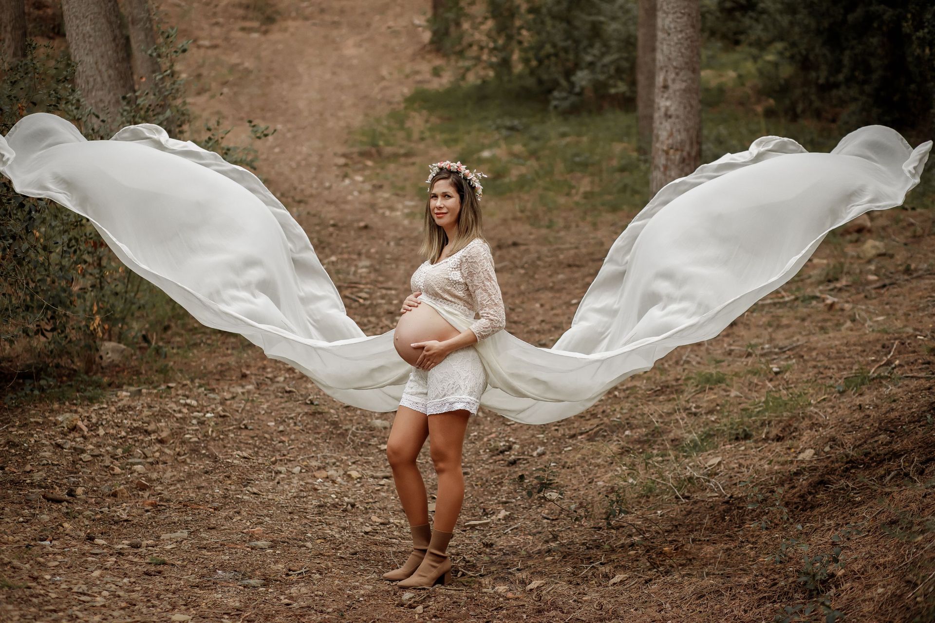 Una mujer embarazada con un vestido blanco con alas está parada en el bosque.