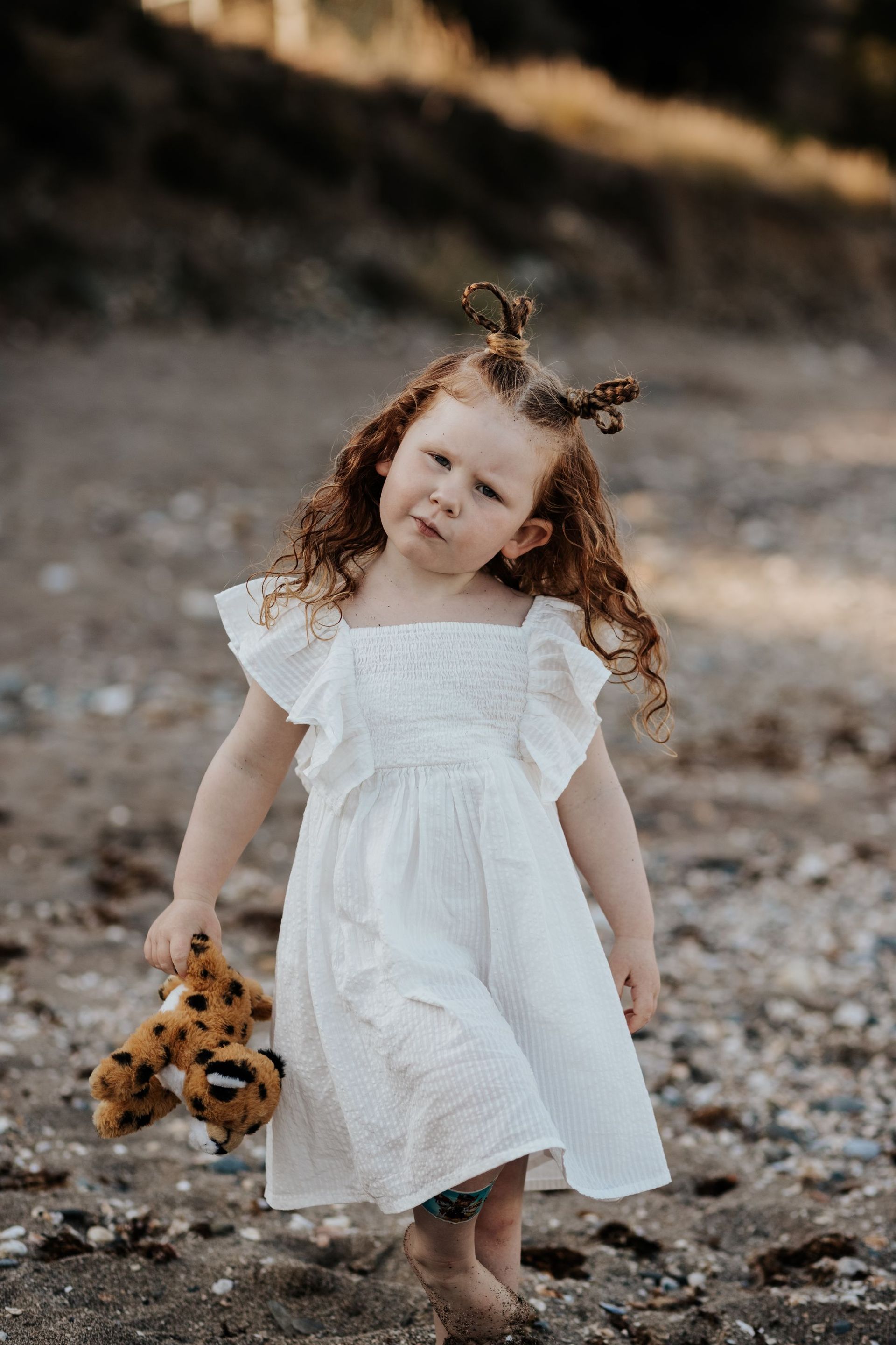 Una niña con un vestido blanco sostiene un osito de peluche en la playa.