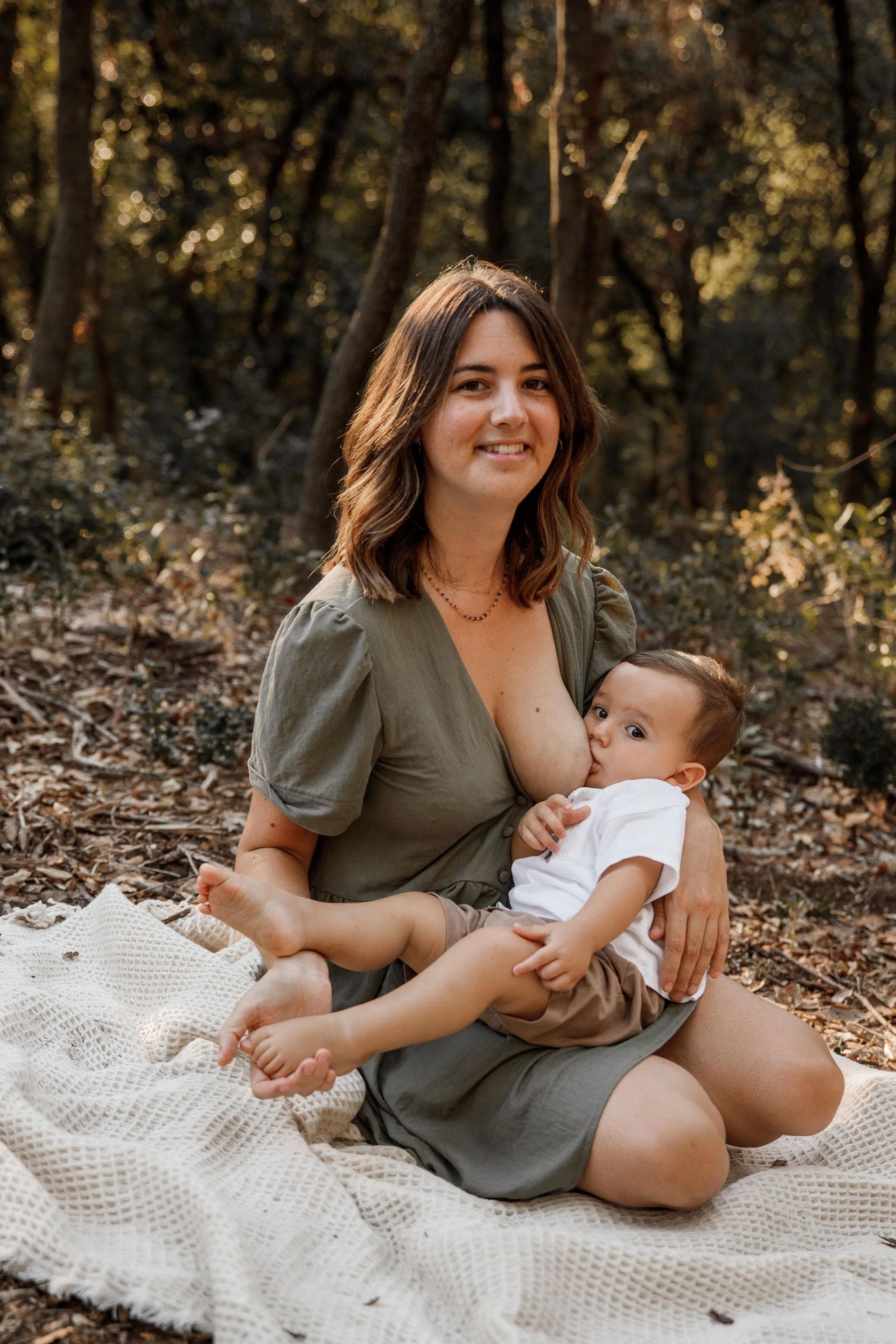 Una mujer está amamantando a su bebé mientras está sentada sobre una manta en el bosque.