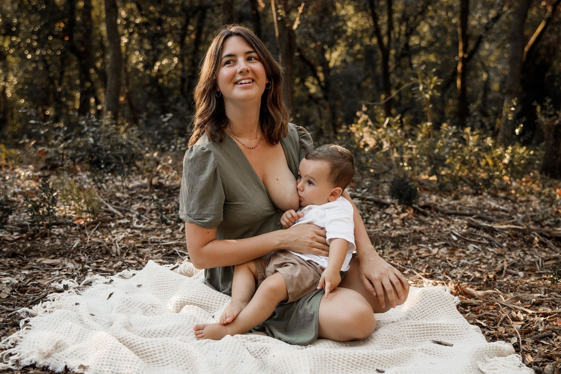 Una mujer está amamantando a su bebé mientras está sentada sobre una manta en el bosque.