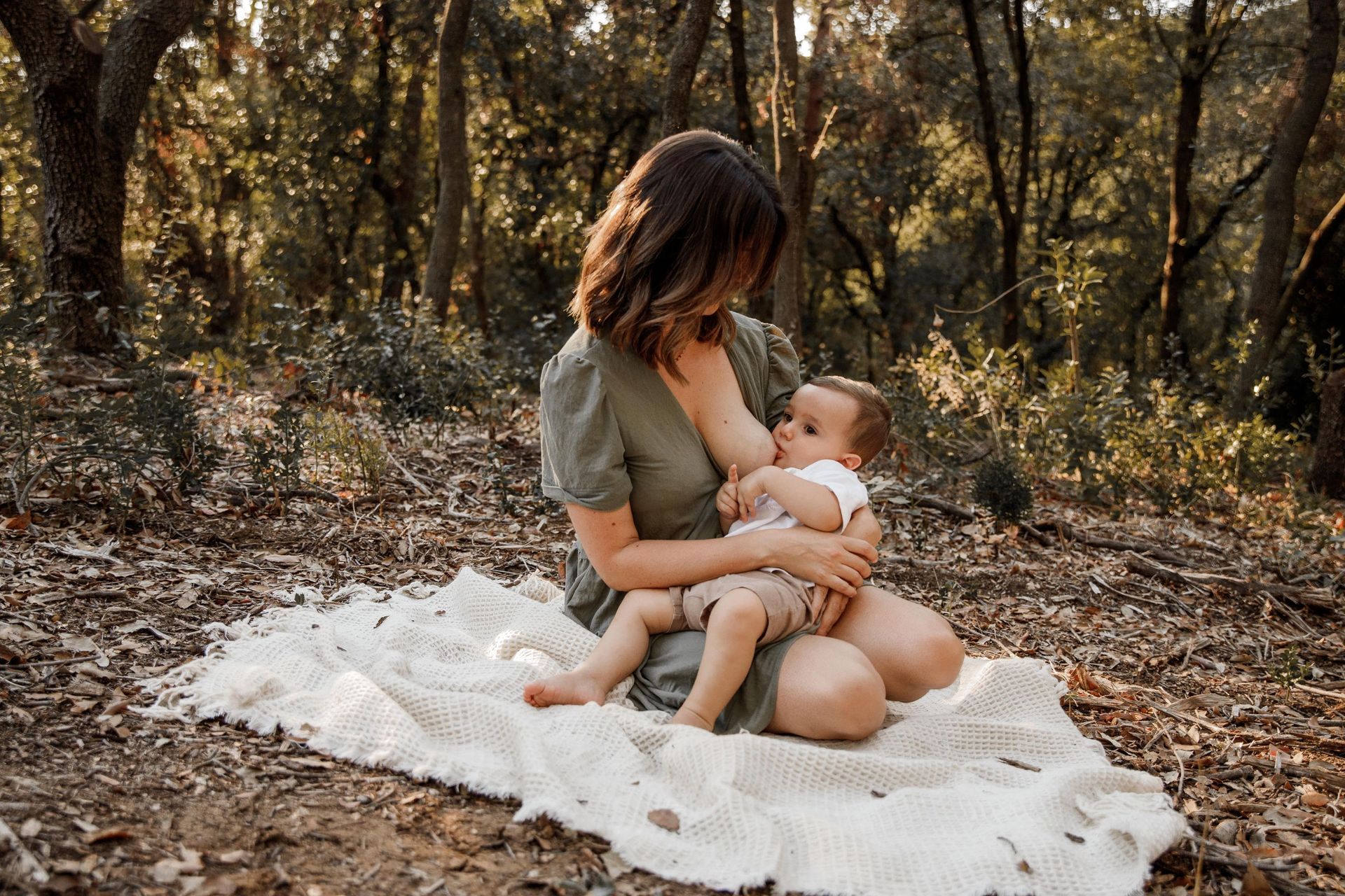 Una mujer está amamantando a su bebé mientras está sentada sobre una manta en el bosque.