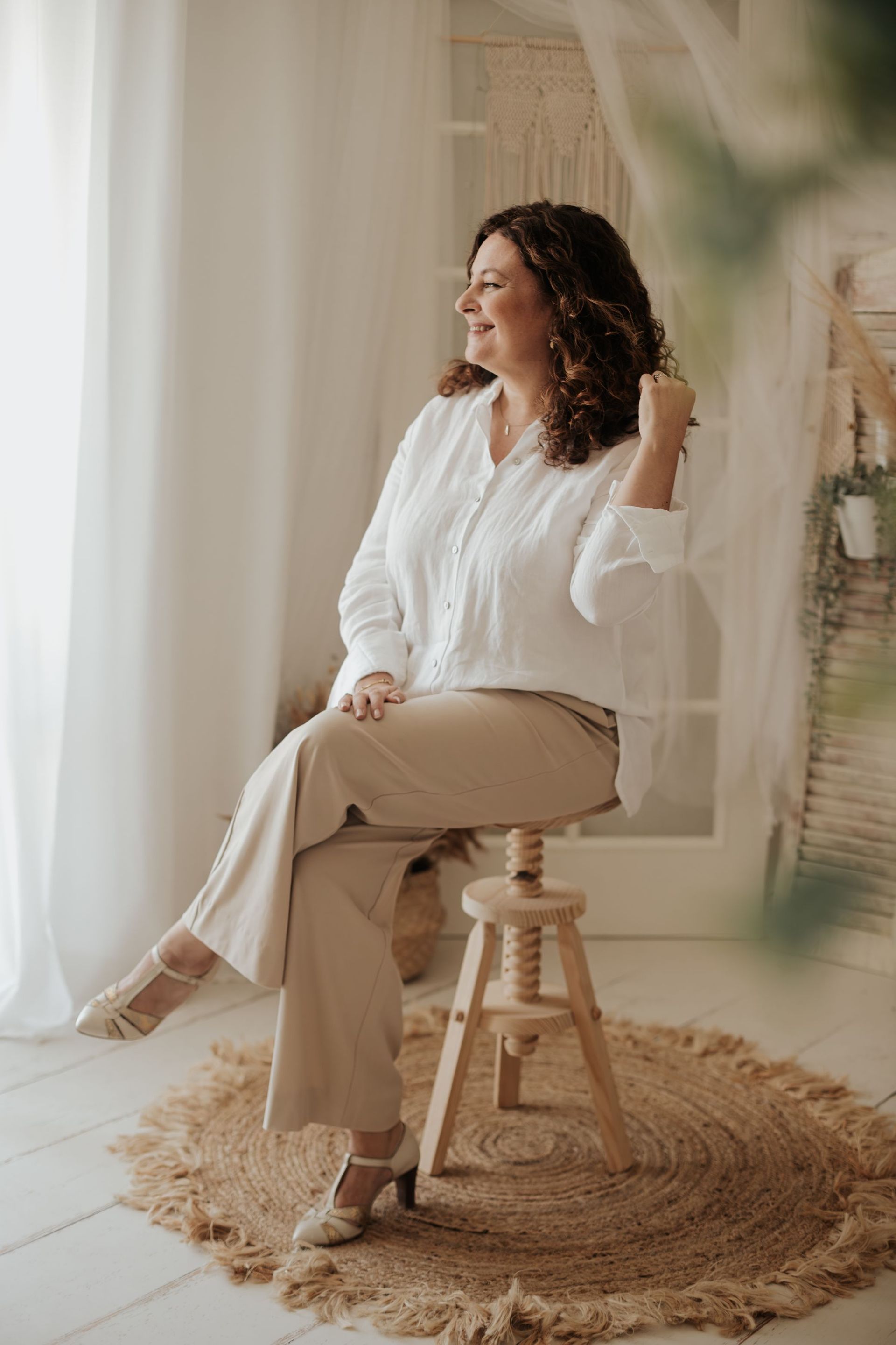 Mujer con blusa blanca y pantalones color canela, sonriendo, sentada en un taburete. La luz del sol entra por una ventana, en un entorno rústico.