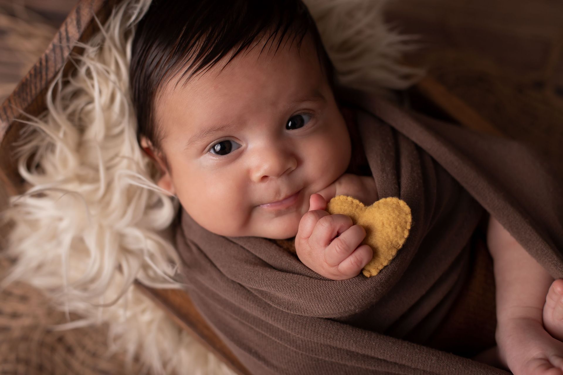 Un bebé recién nacido envuelto en una manta marrón sostiene un corazón en su mano.