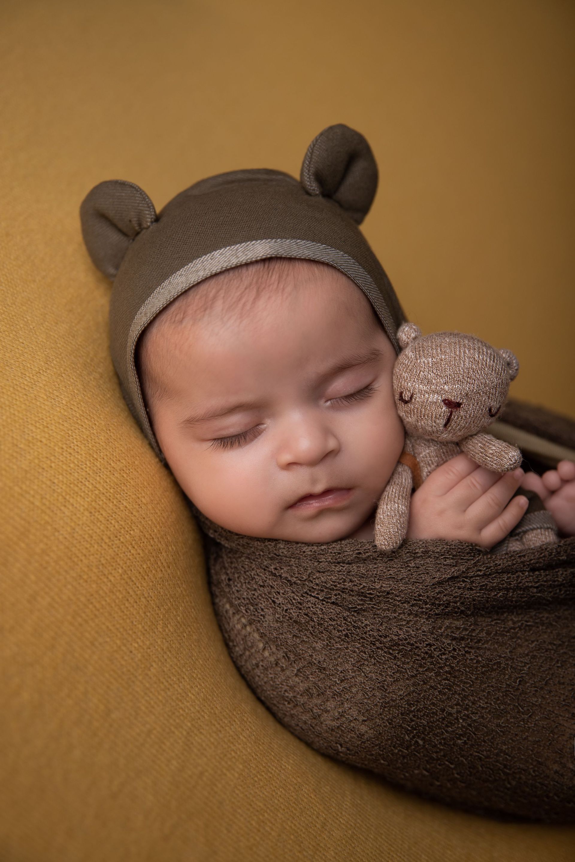 Un bebé recién nacido envuelto en una manta marrón sostiene un osito de peluche.