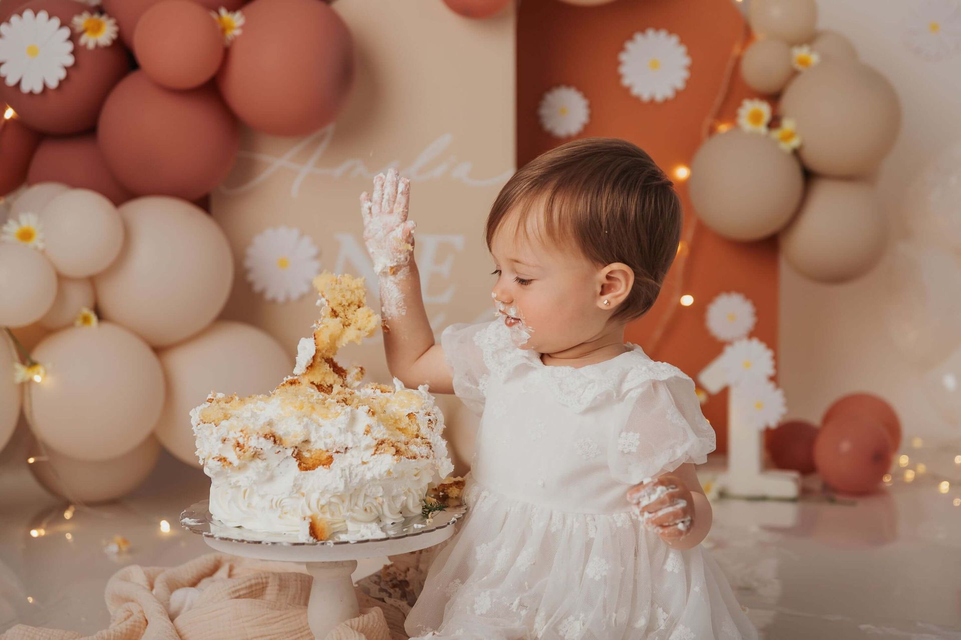 Una niña pequeña con un vestido blanco destroza un pastel, con la cara y las manos cubiertas de glaseado, en su sesión Smash Cake en Málaga