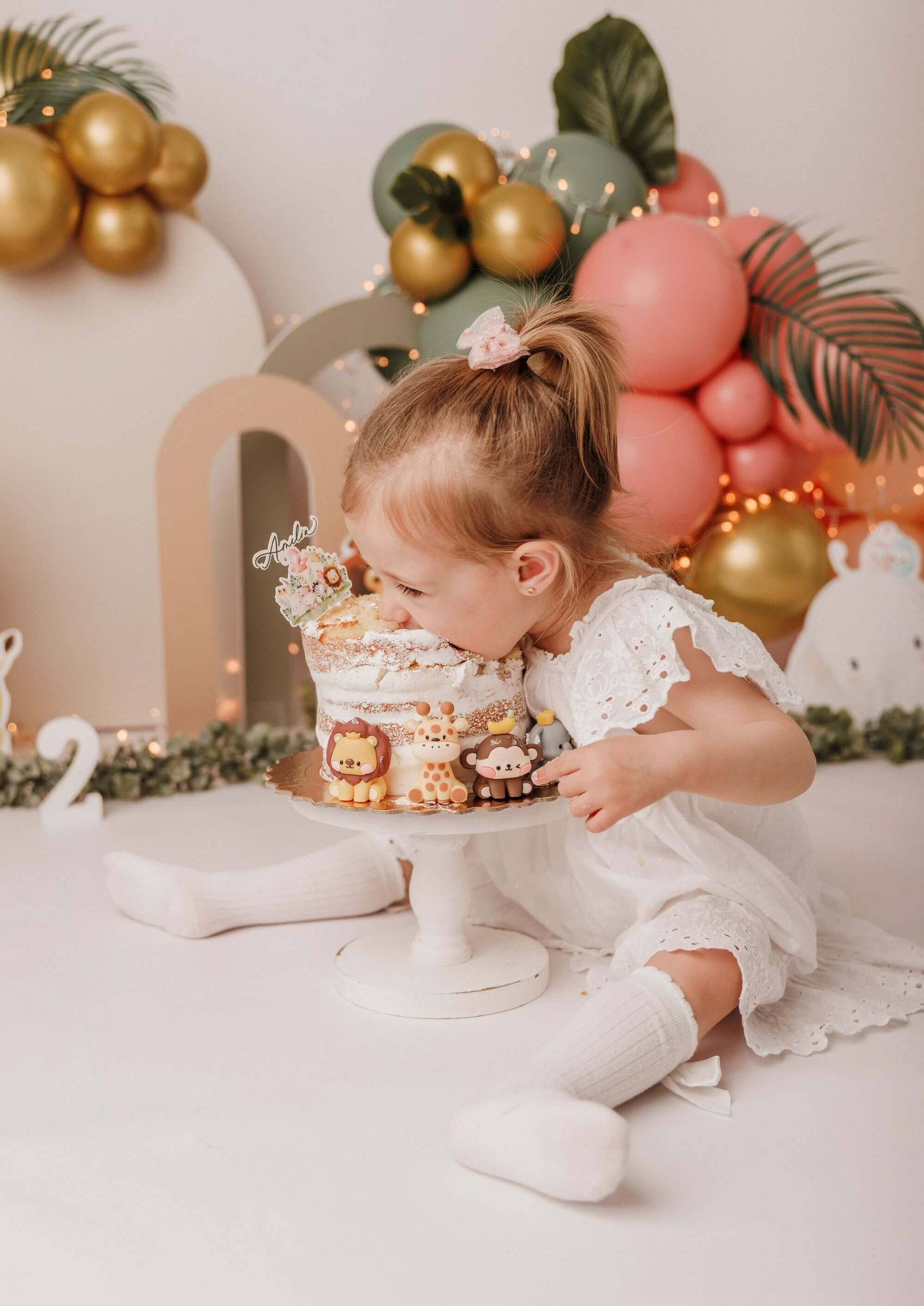 Una niña con un lazo rosa en el pelo, se sienta delante de un arco de globos de colores y come un pastel en su sesión Smash Cake en Málaga