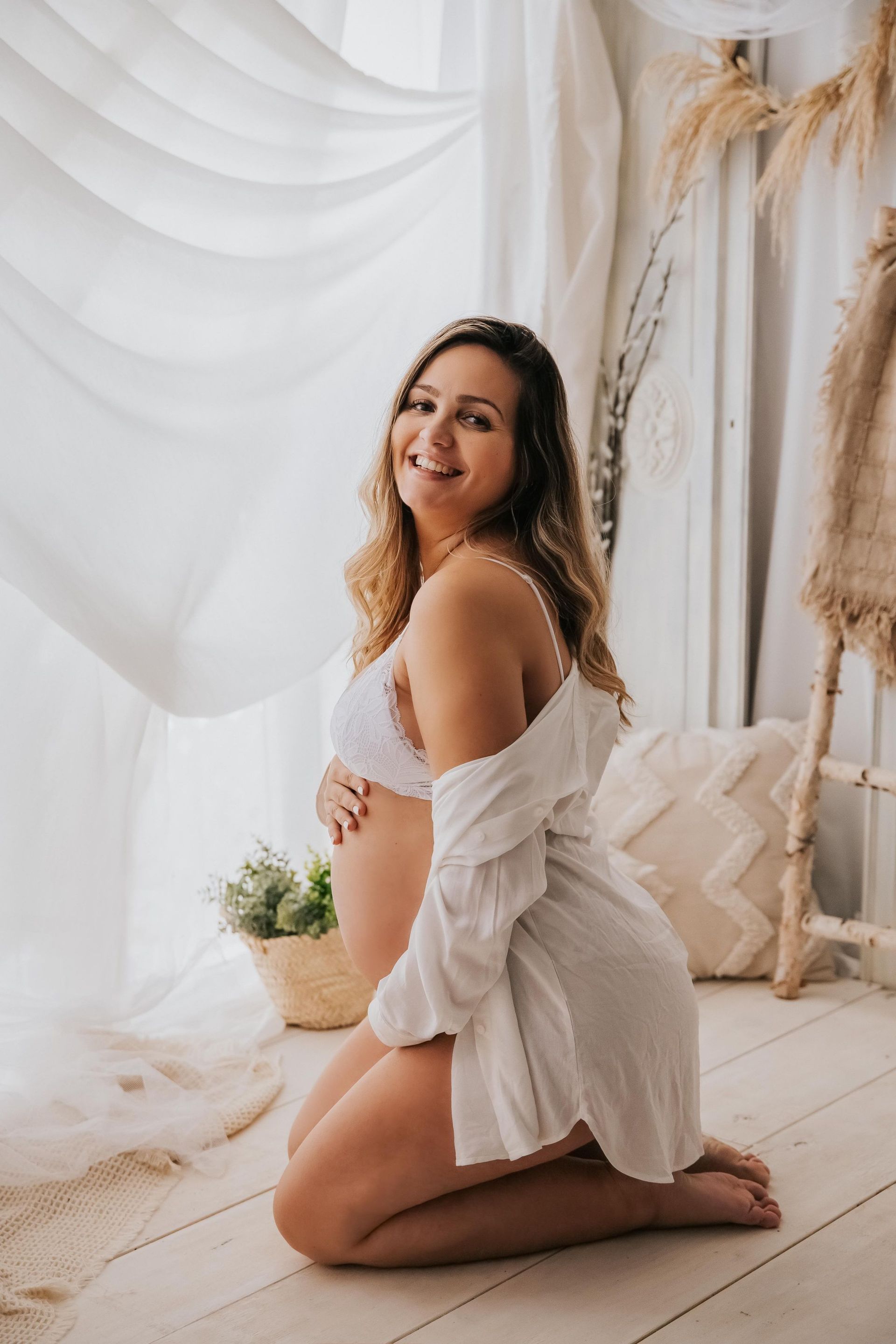 Una mujer embarazada con un vestido blanco está arrodillada frente a una ventana.