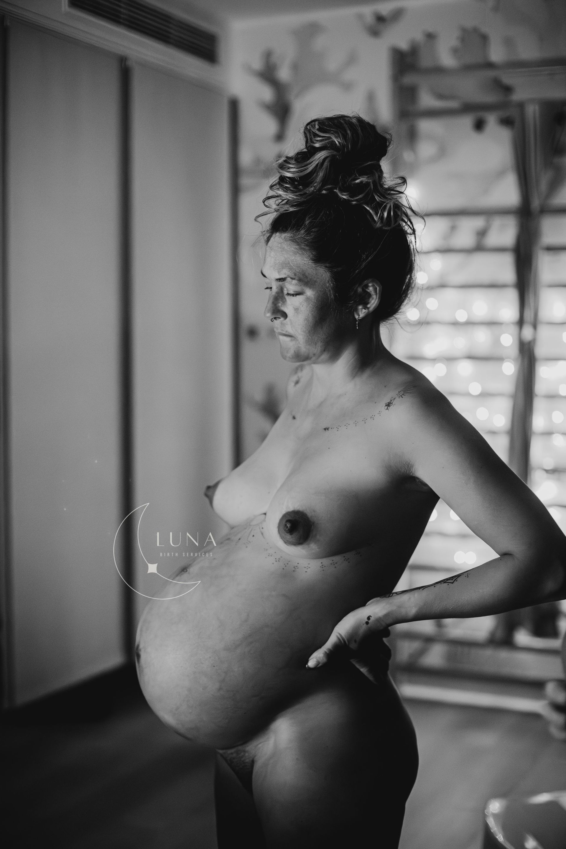 Una mujer embarazada está desnuda, de perfil, en una habitación. Tiene una barriga grande y el pelo recogido. Salpica agua.