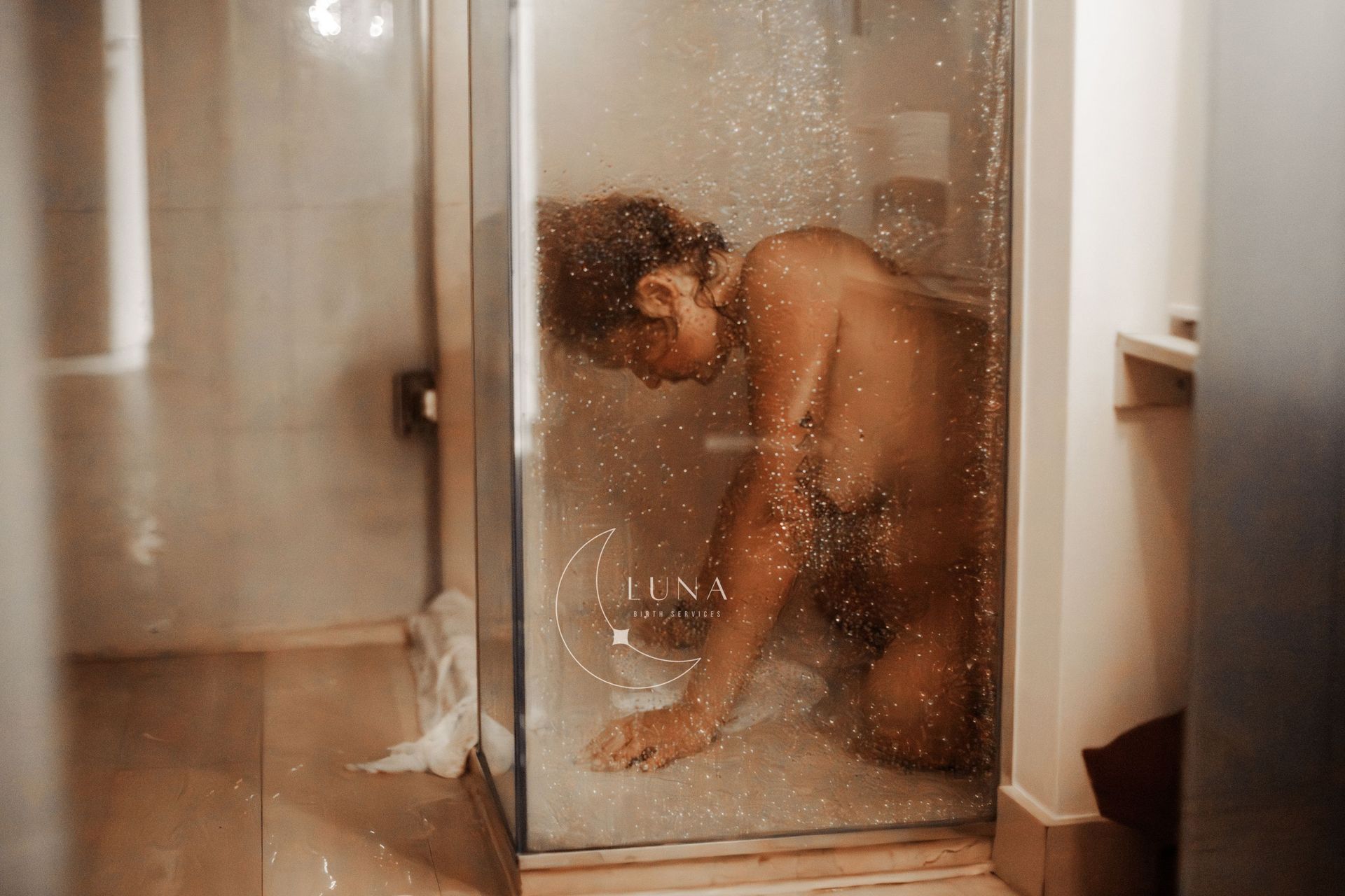 Persona arrodillada en una ducha, oscurecida por gotas de agua en el cristal, tono neutro.