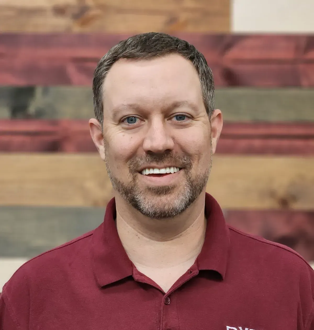 A man with a beard wearing a maroon shirt is smiling in front of a wooden wall.