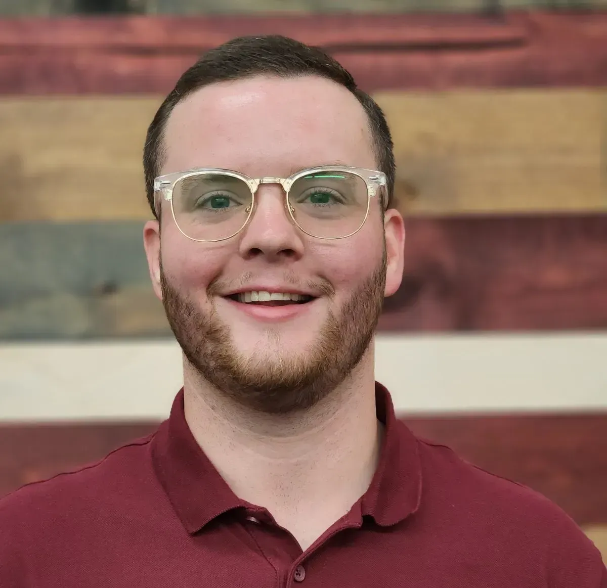 A man wearing glasses and a maroon shirt is smiling in front of a wooden wall.