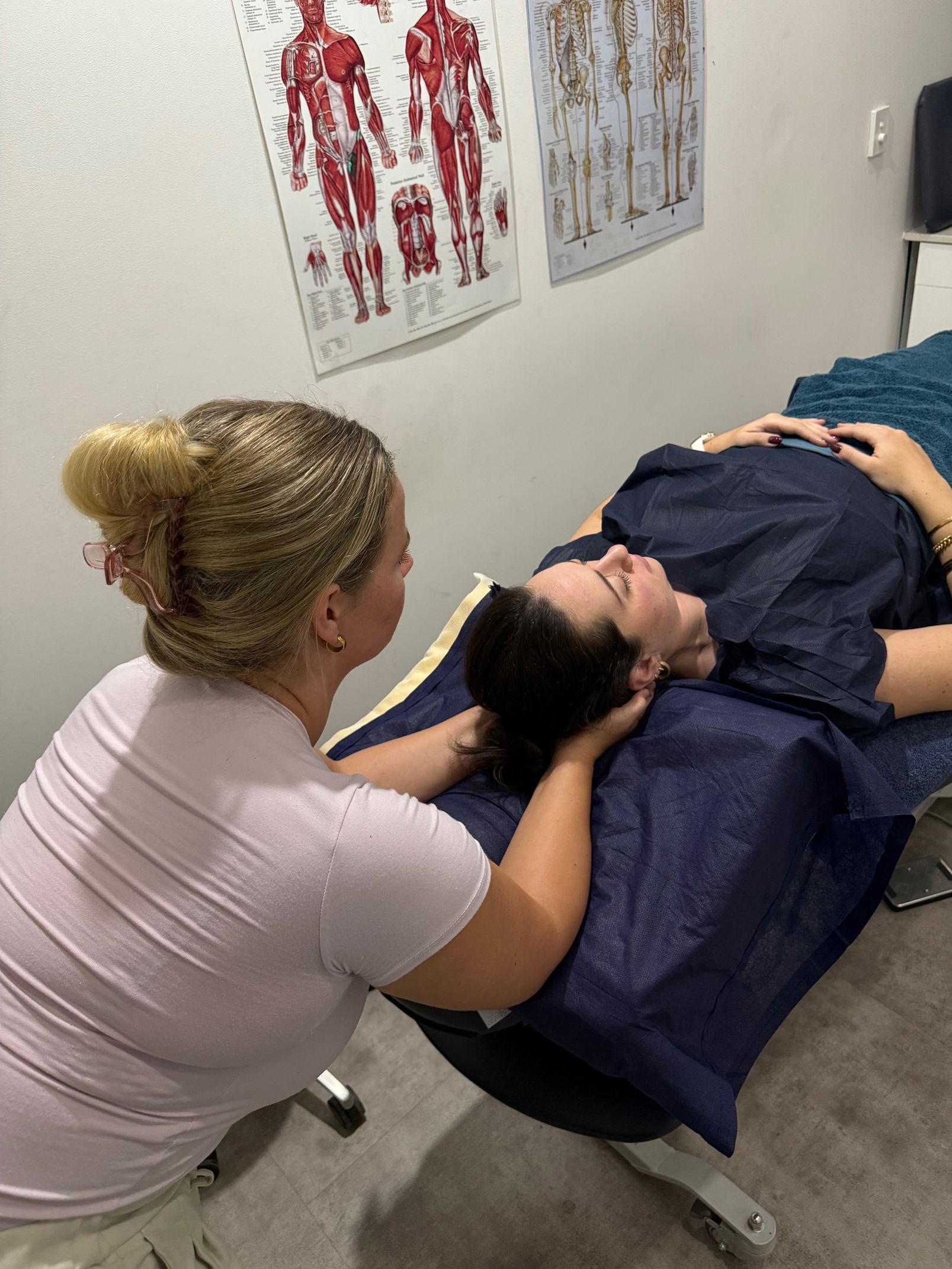 Woman massaging another woman's neck on a massage table. Anatomical charts on wall — Spinal Symmetry In Dee Why, NSW