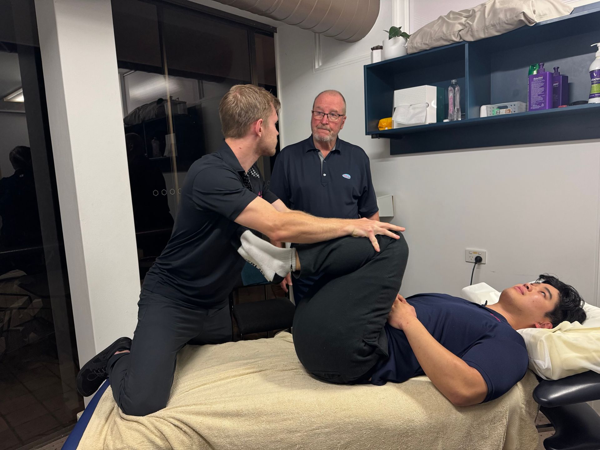 Two men are standing around a chiropractic bed while moving another mans legs laying down — Spinal Symmetry In Salamander Bay, NSW