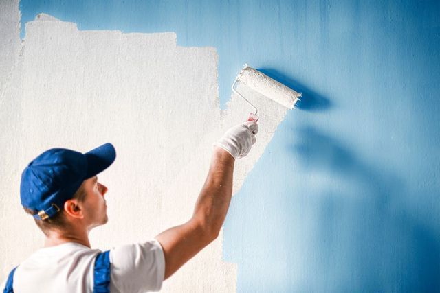 Painter painting a blue wall white with a roller brush in a blue and white uniform