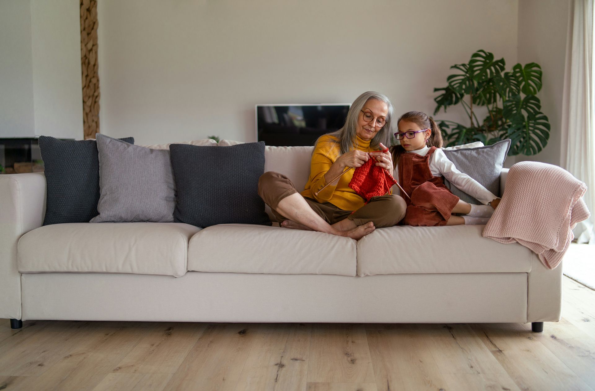 Woman knitting with a child, seated on a white couch. Bright living room.