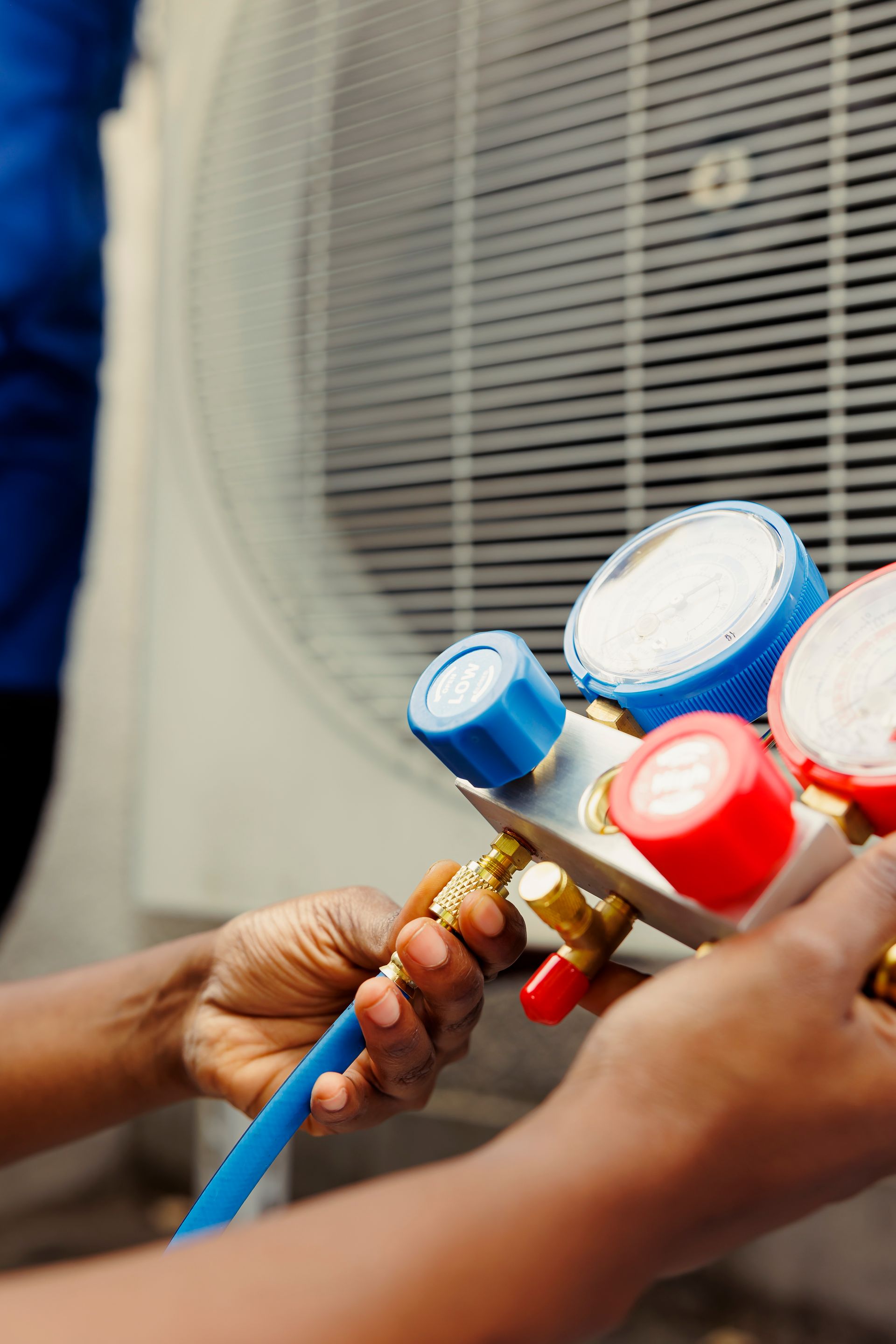 Hands connecting gauges to an air conditioning unit. Blue and red gauges, white unit.