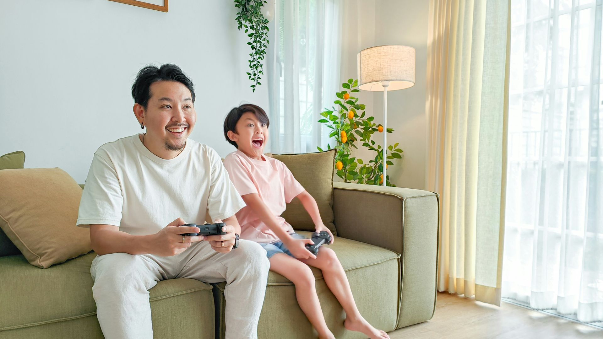 Man and child playing video games, sitting on a sofa in a living room; both are smiling and holding controllers.