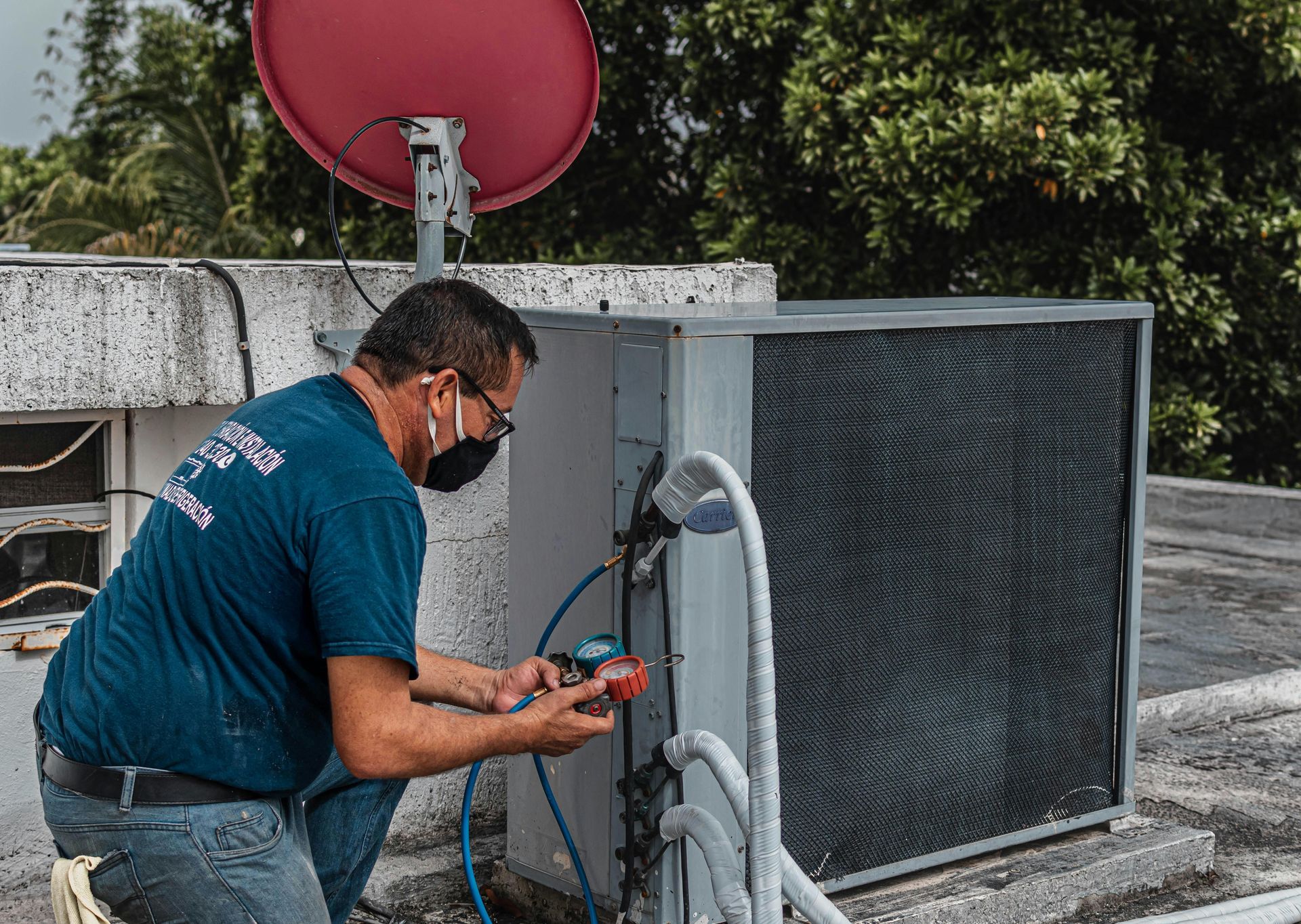 HVAC technician in a face mask repairs an AC unit on a rooftop. A red satellite dish is visible.