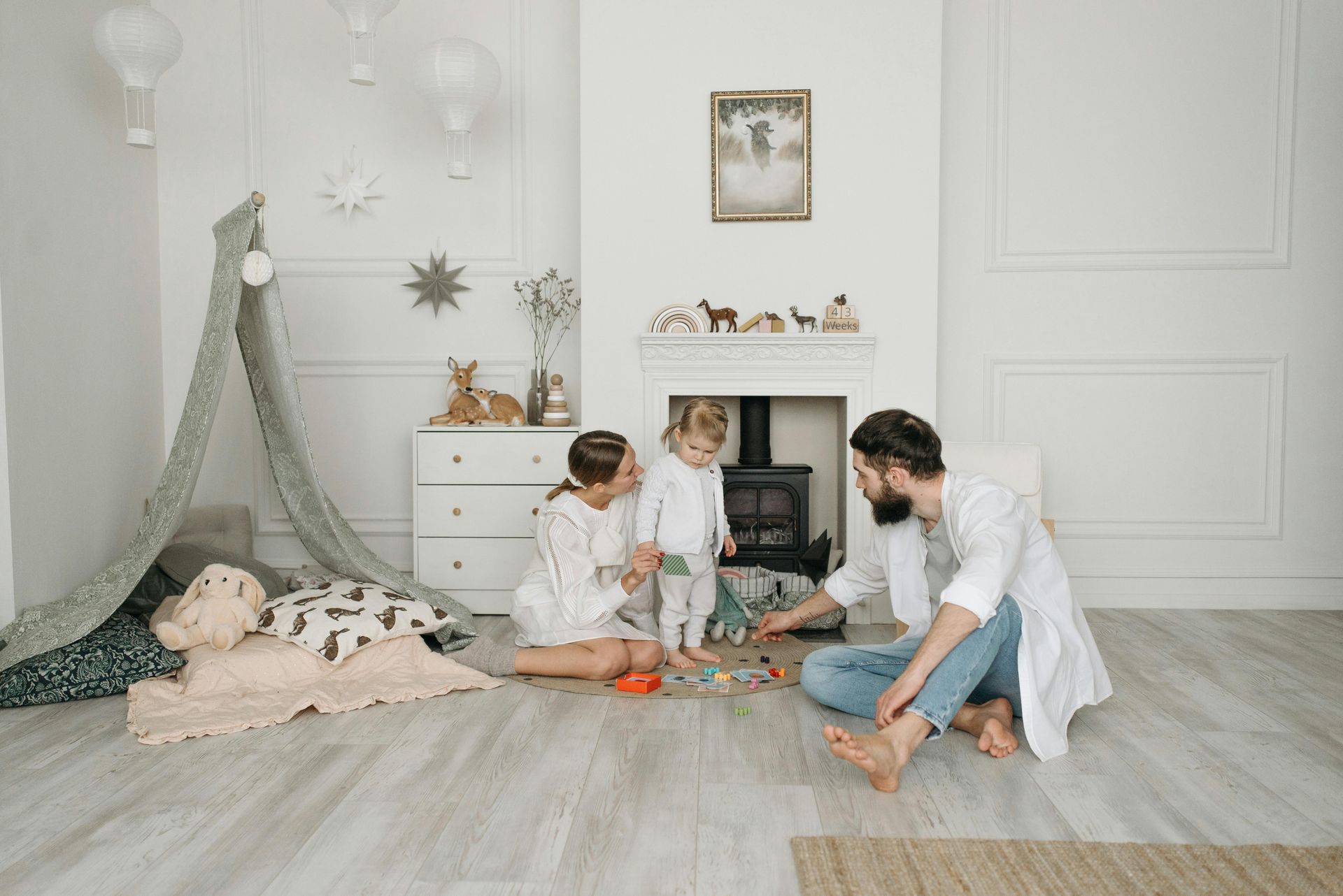 Family playing on floor in a cozy white-toned room. Child in middle, parents seated.
