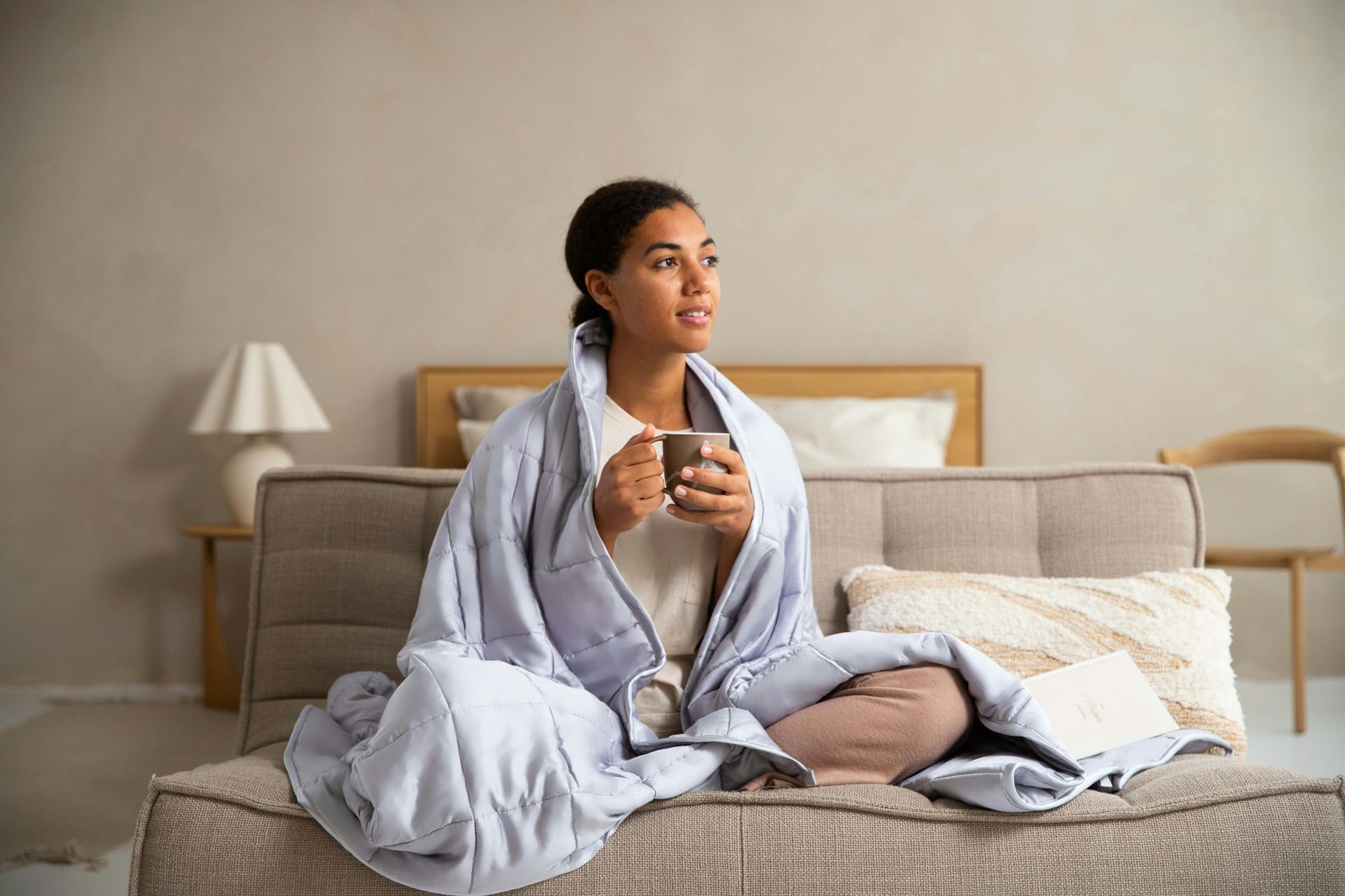 Person wrapped in a blue blanket sits on a sofa, holding a mug and looking toward the window.