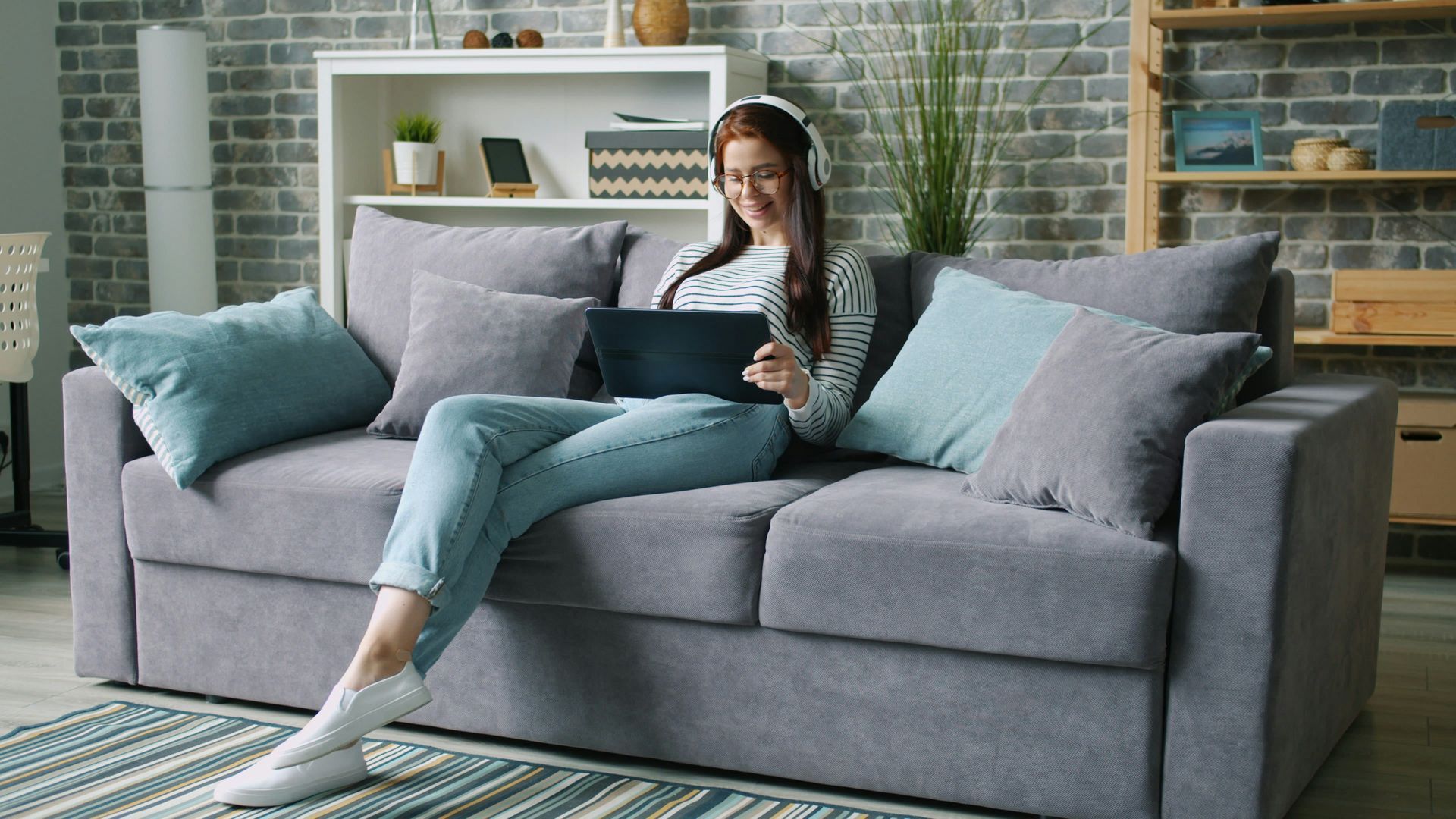 Woman wearing headphones, using a laptop, sitting on a gray couch in a living room.