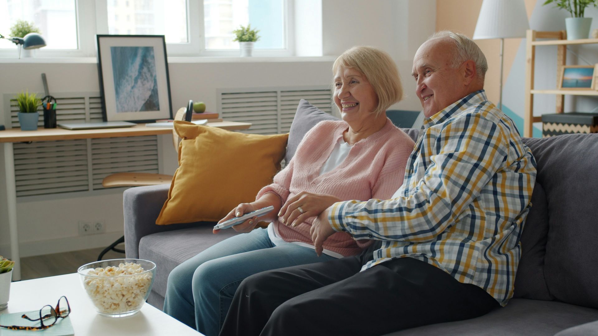 Senior couple laughs while watching TV on a couch. Living room with popcorn and remote.
