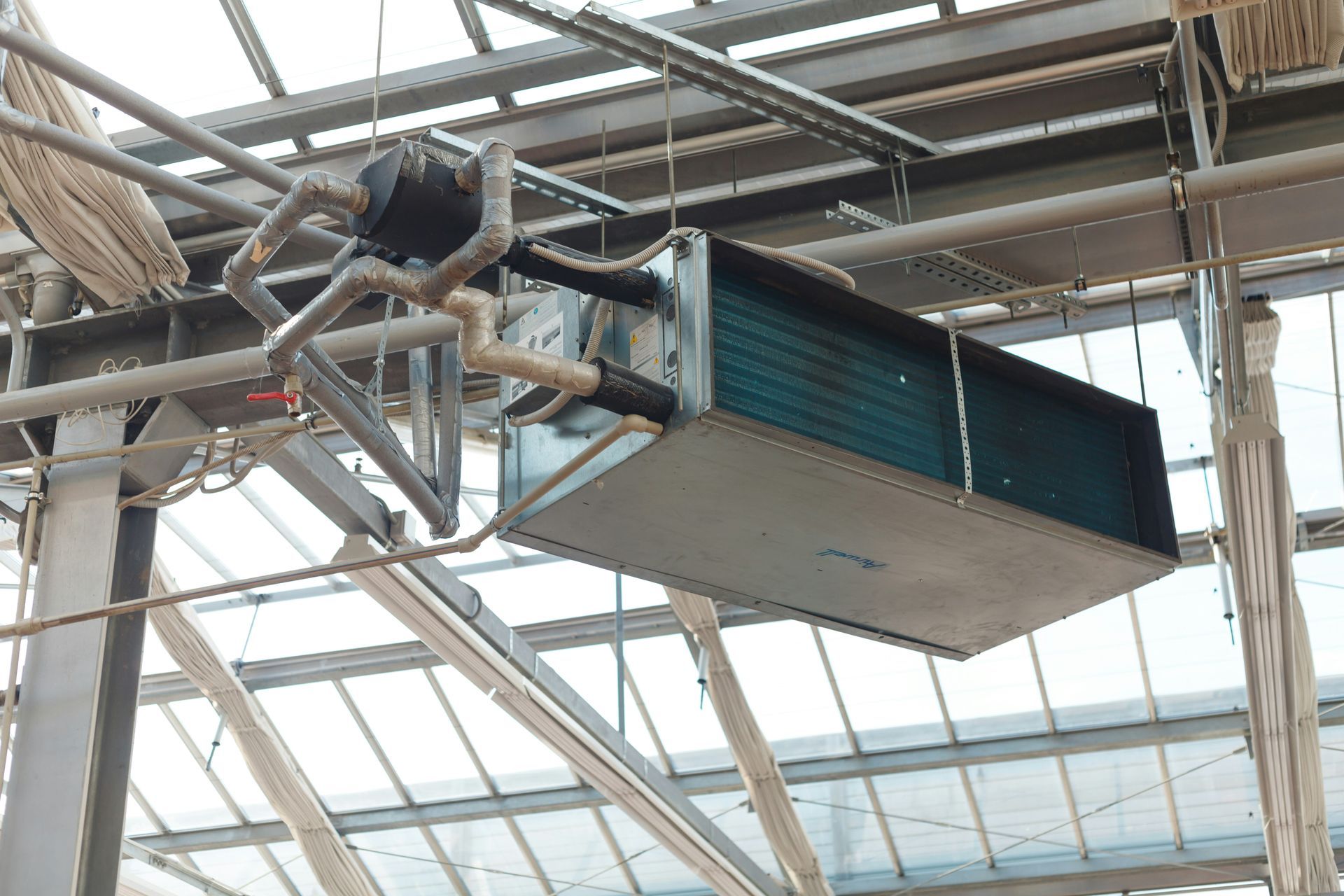 HVAC unit suspended from greenhouse ceiling, with pipes and a fan.