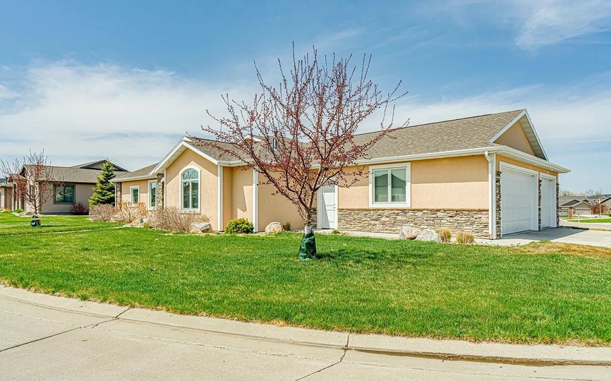A tan suburban house with a stone-accented facade, a two-car garage, and a bare tree in the front lawn under a blue sky.