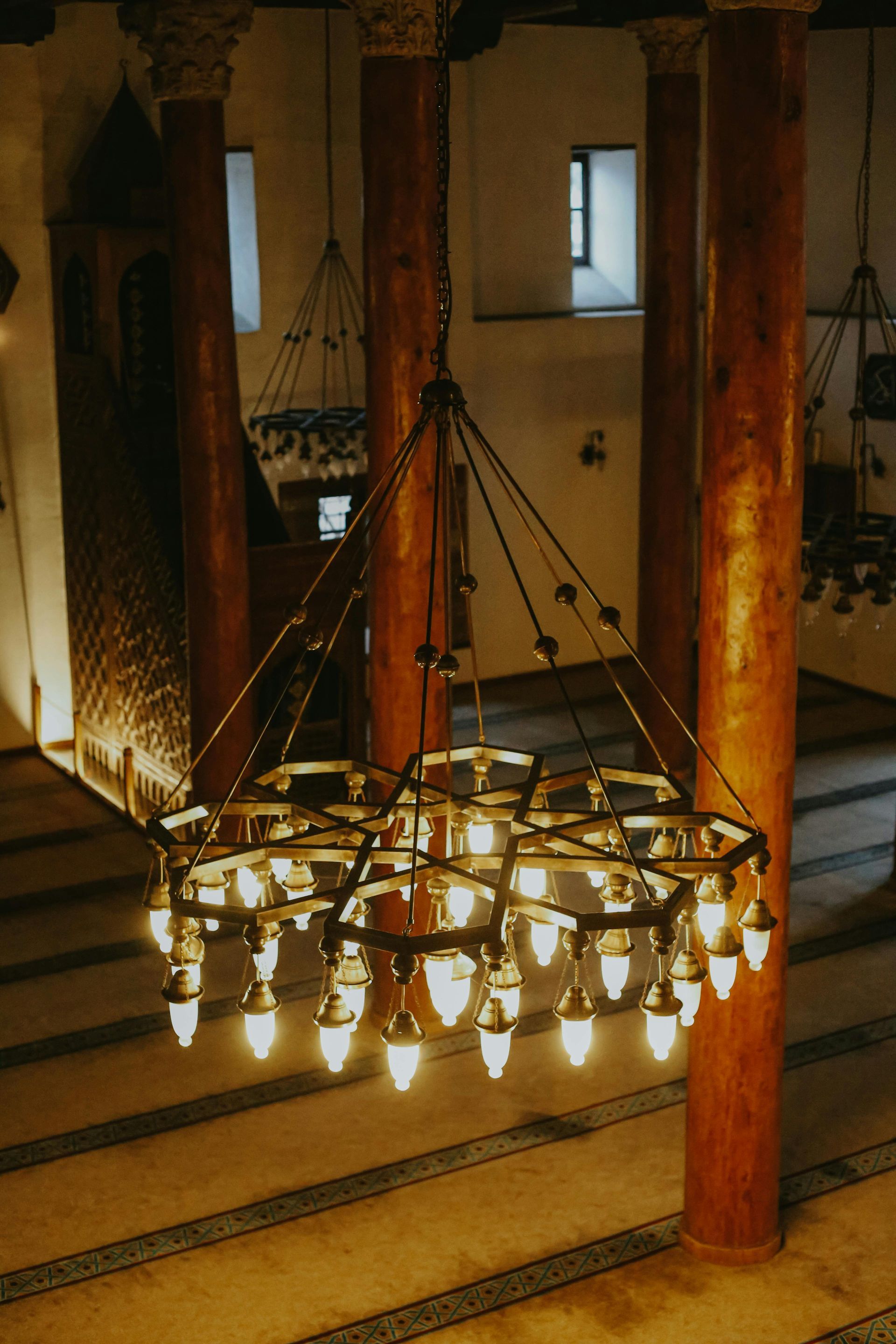 Inside of a mosque, a large chandelier hangs. Columns and prayer rugs are visible.