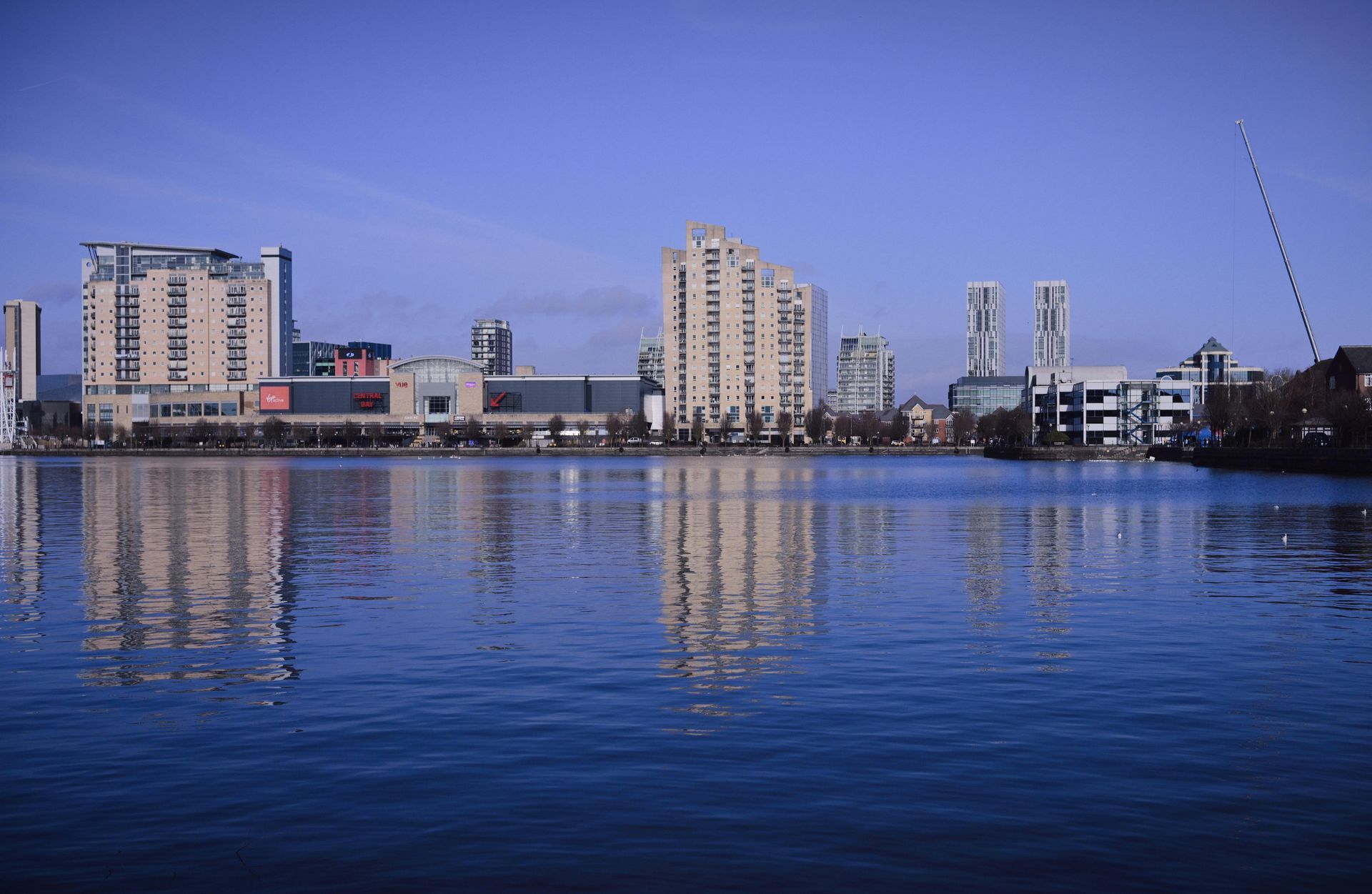 City skyline reflecting in a lake, with blue water and sky. Buildings include high-rises.