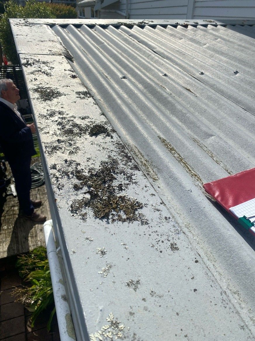 A man stands beside a house inspecting a light gray corrugated metal roof with scattered debris and 