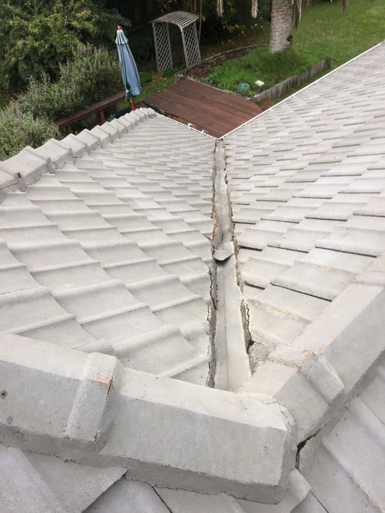 Overhead view of a light gray tiled roof showing a central valley with visible cracks and separation in the mortar, with a backyard deck and garden visible below.