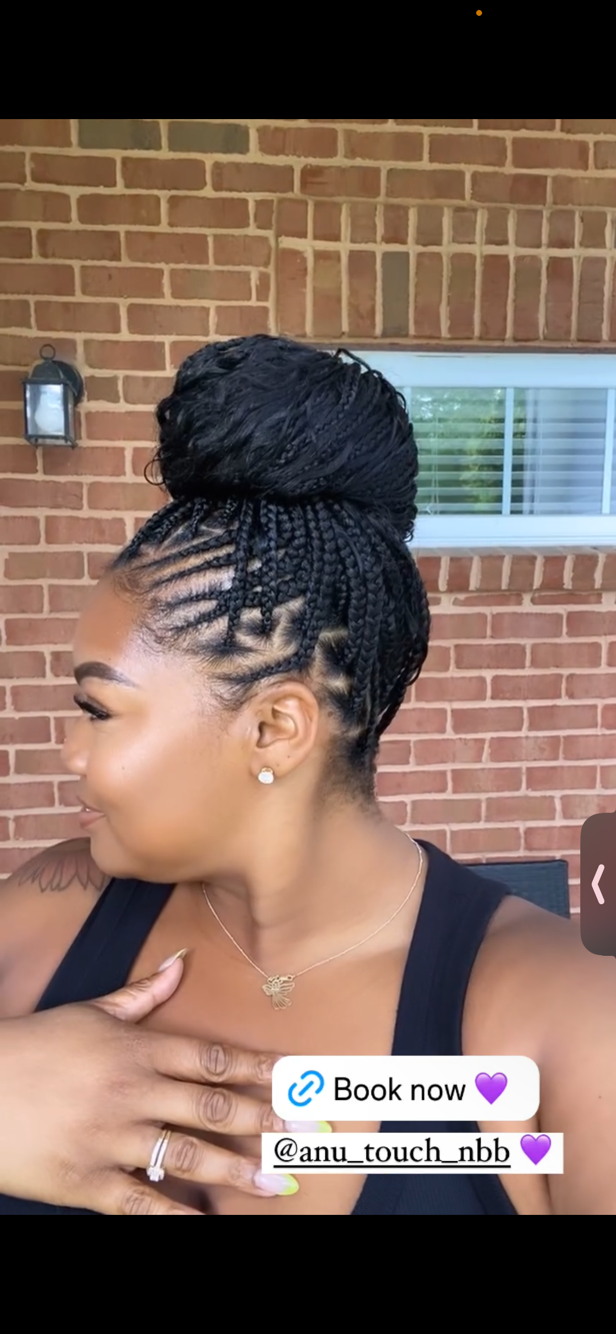 Woman with braided hair in a bun. She has a necklace and is wearing a black top, possibly outside.