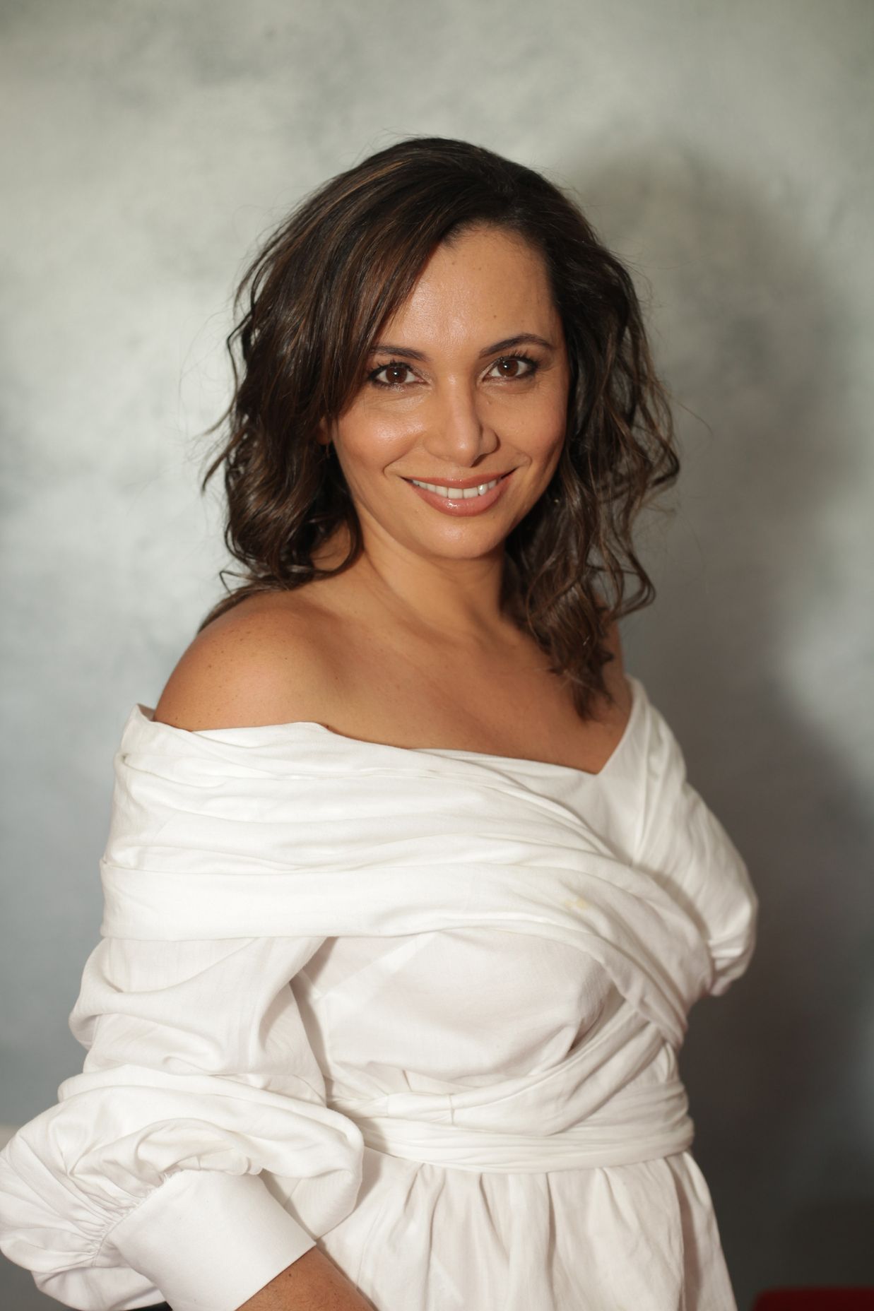 Woman wearing a white off-the-shoulder top, smiling. Against a light, textured background.