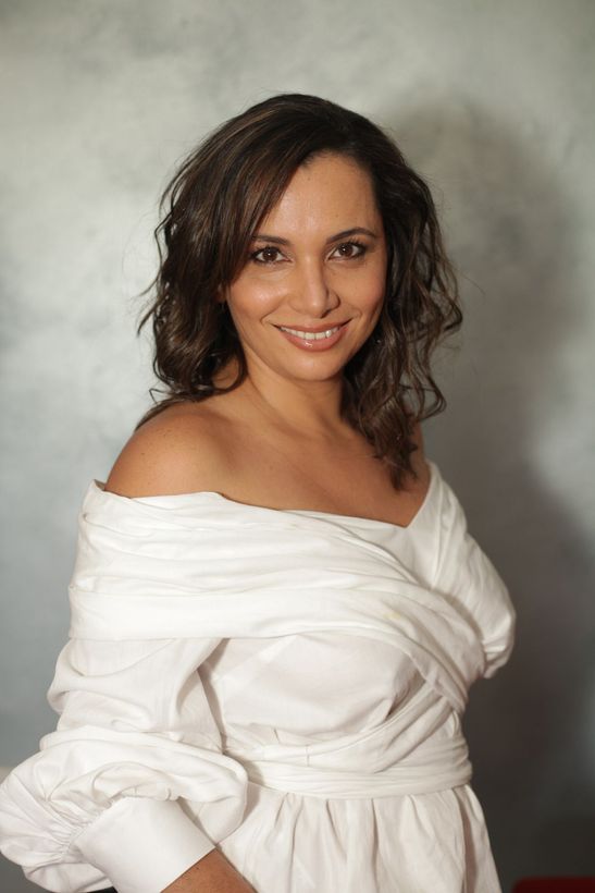 Woman wearing a white off-the-shoulder top, smiling. Against a light, textured background.