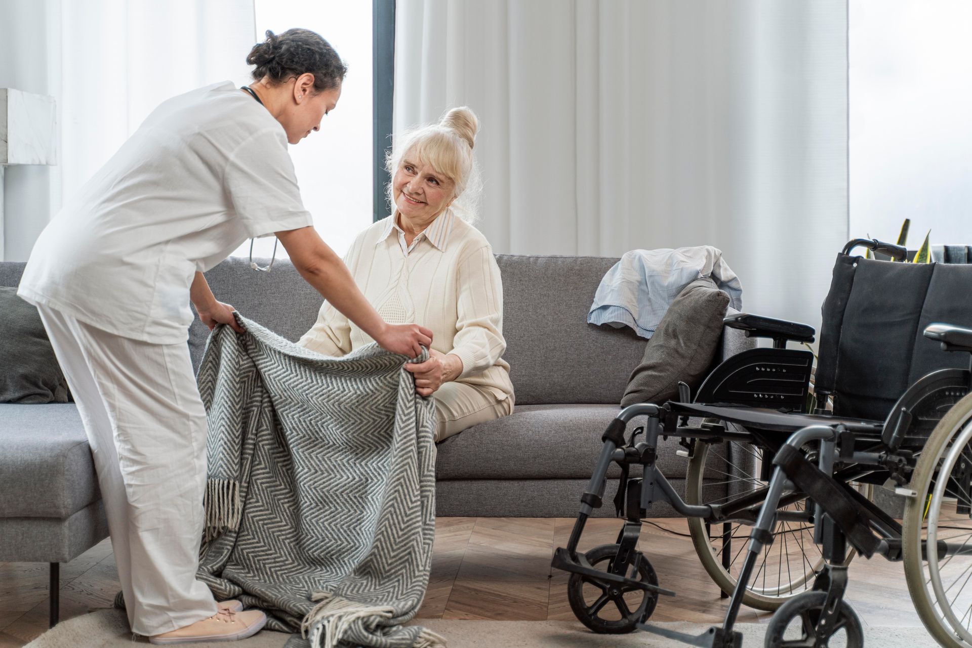 A nurse is putting a blanket on an elderly woman sitting on a couch.