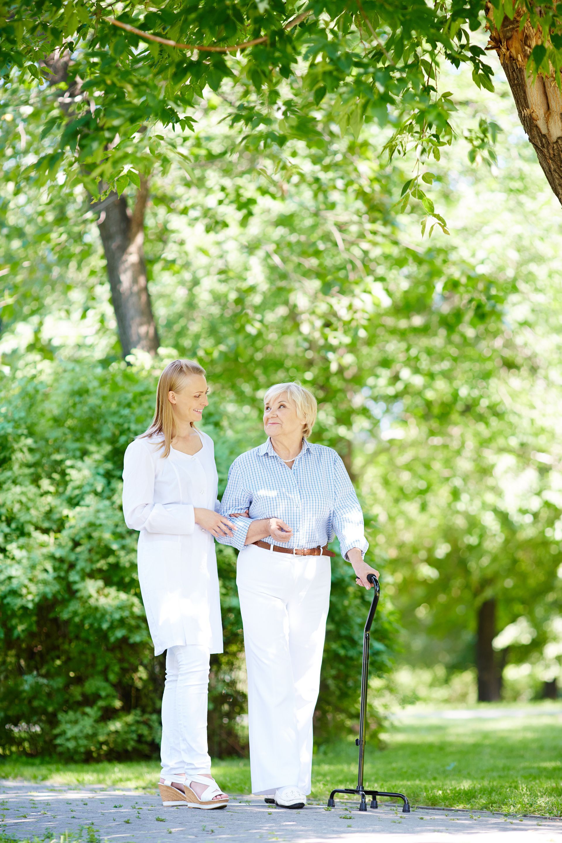 A young woman is helping an older woman with a cane walk in a park.