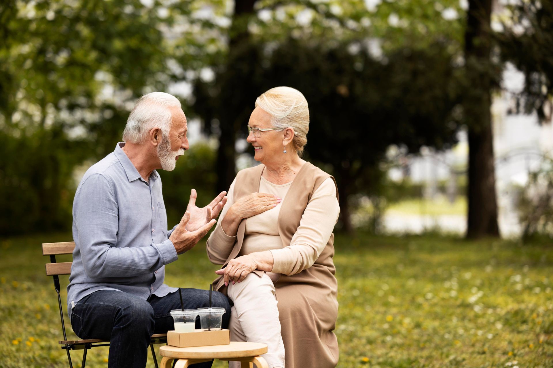 An elderly couple is sitting on a bench in a park talking to each other.