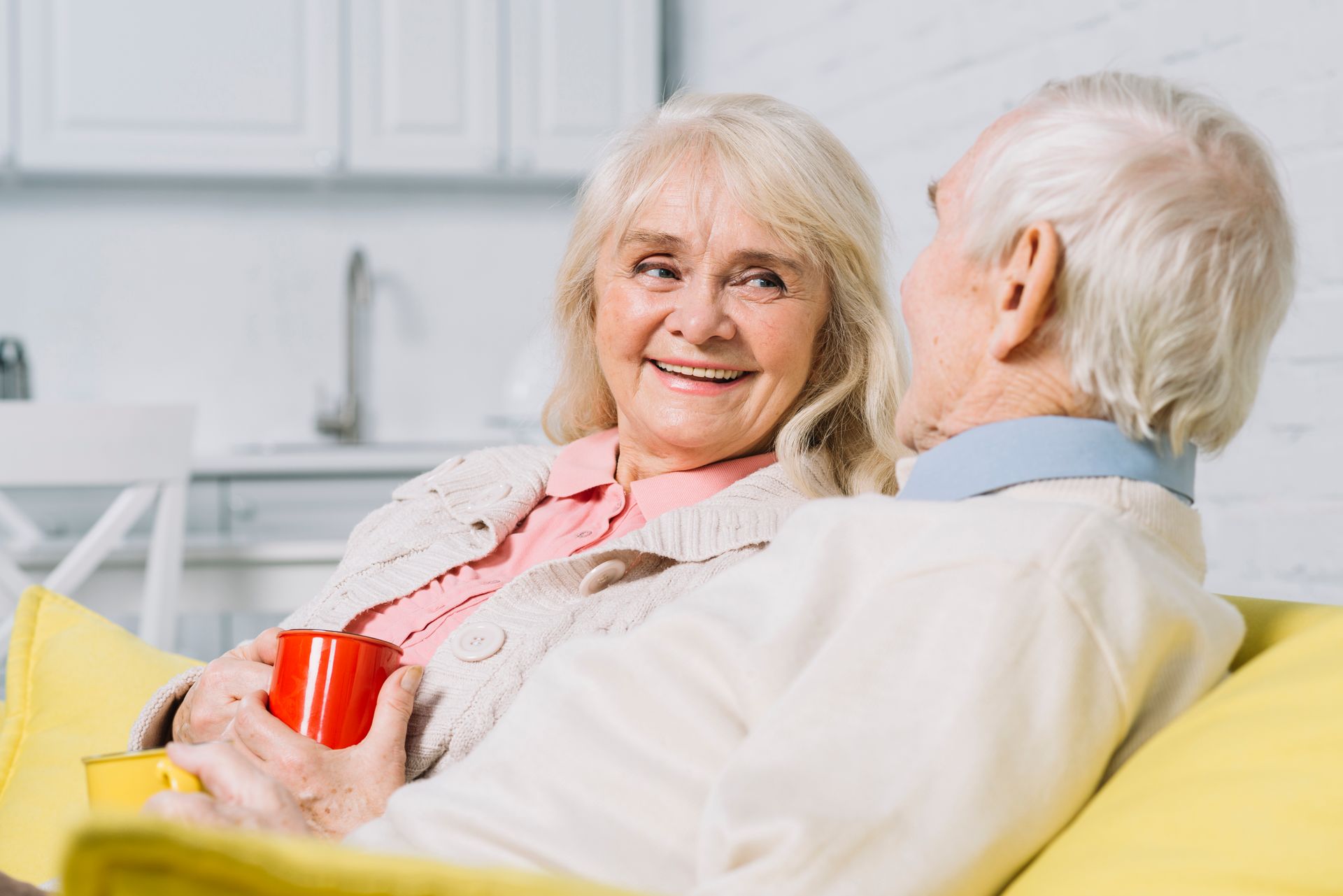 An elderly couple is sitting on a yellow couch looking at each other.