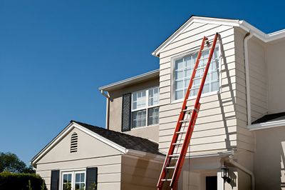 Ladder leaning against a two-story house, box being carried up, bright blue sky.