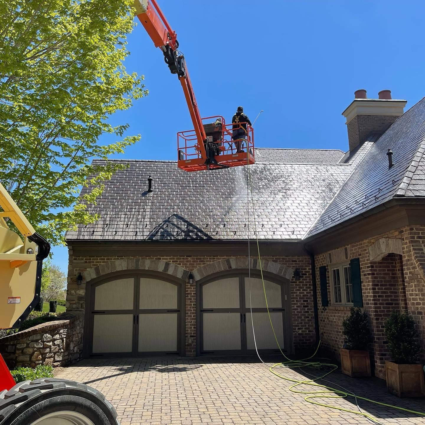 Workers on a roof using an orange boom lift to repair gray shingles on a brick house