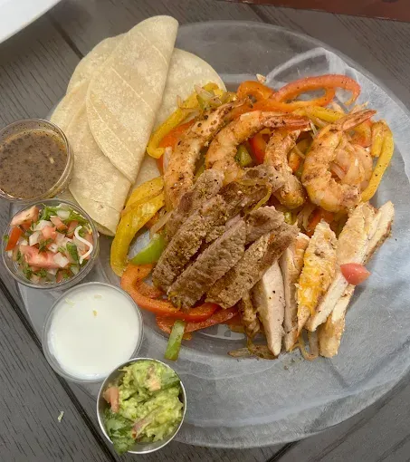 Fajita platter: grilled steak, chicken, shrimp, peppers, tortillas, and sides like salsa and guacamole.